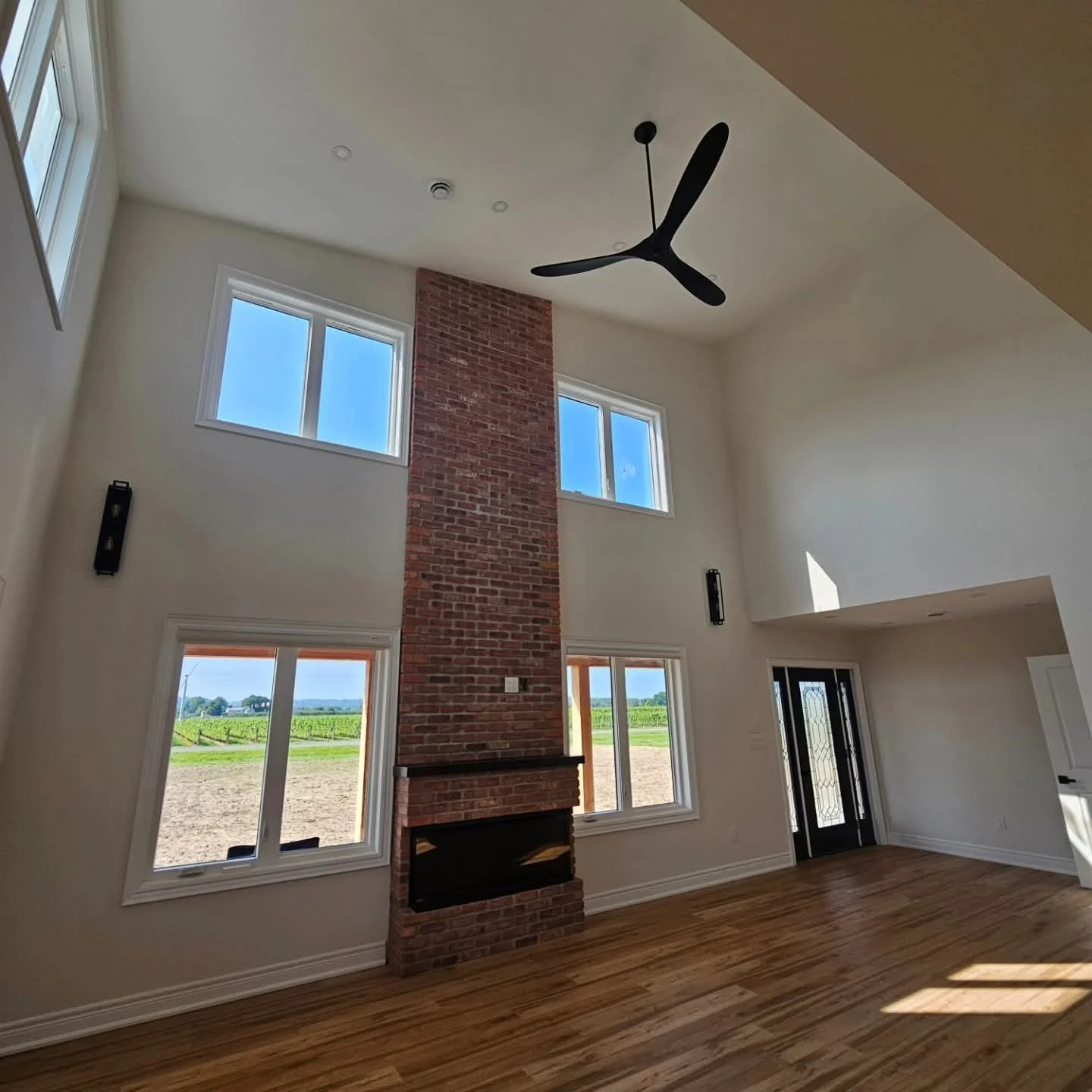 Interior of a spacious living room with high ceiling, white walls, large windows showing fields outside, a brick fireplace, and a ceiling fan.