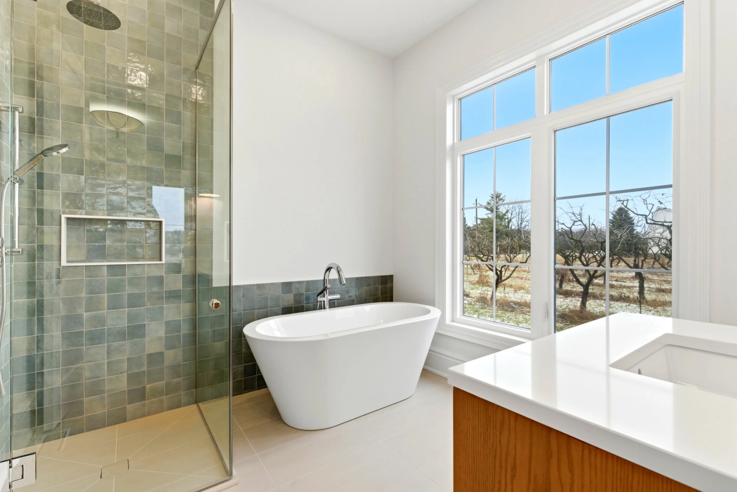 Modern bathroom with a glass-enclosed shower, a white freestanding bathtub, a large window showing trees outside, and a white countertop with a wooden base.