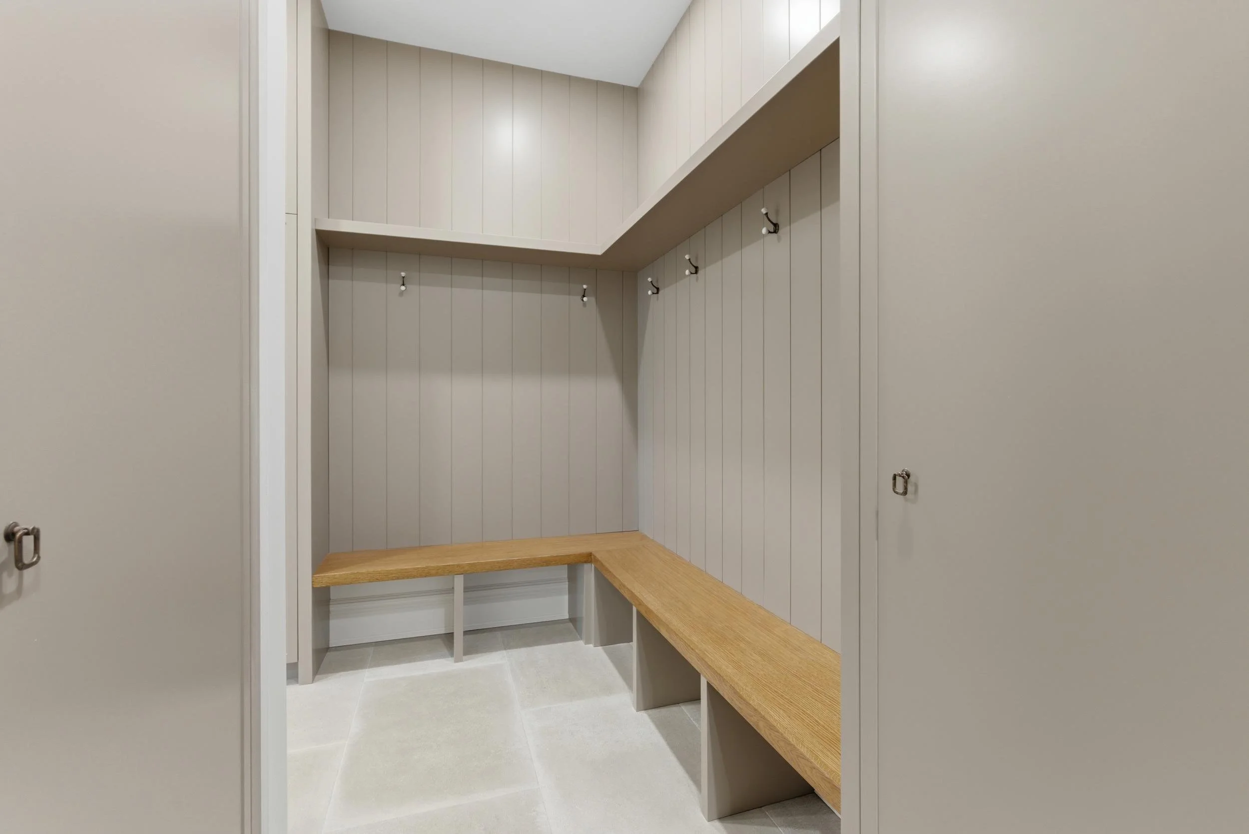 Empty laundry or mudroom closet with beige vertical paneling, wooden benches, hooks, and open shelves.