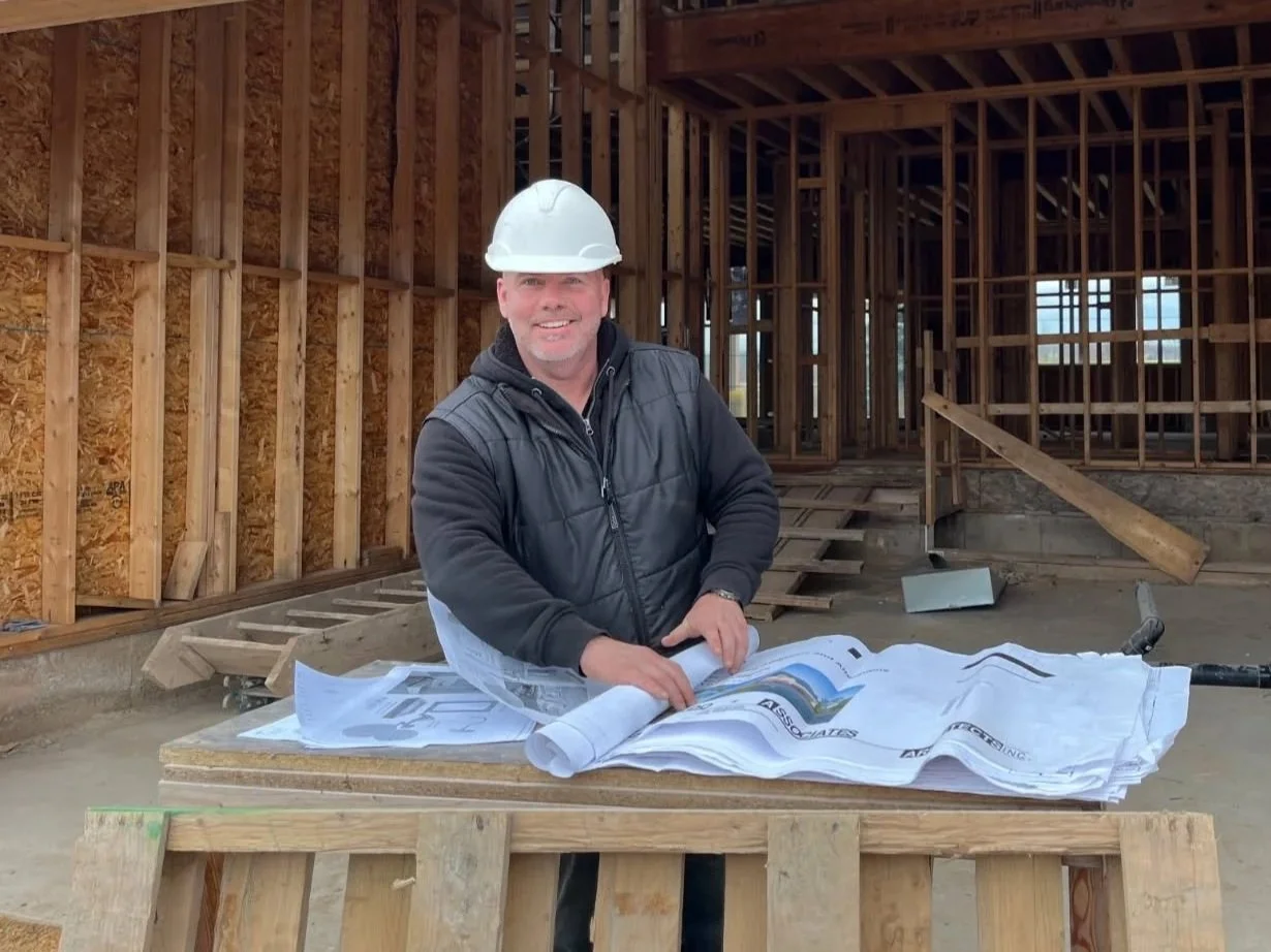 A man wearing a white construction helmet and a black vest smiling while looking at blueprints on a wooden table inside a building under construction.