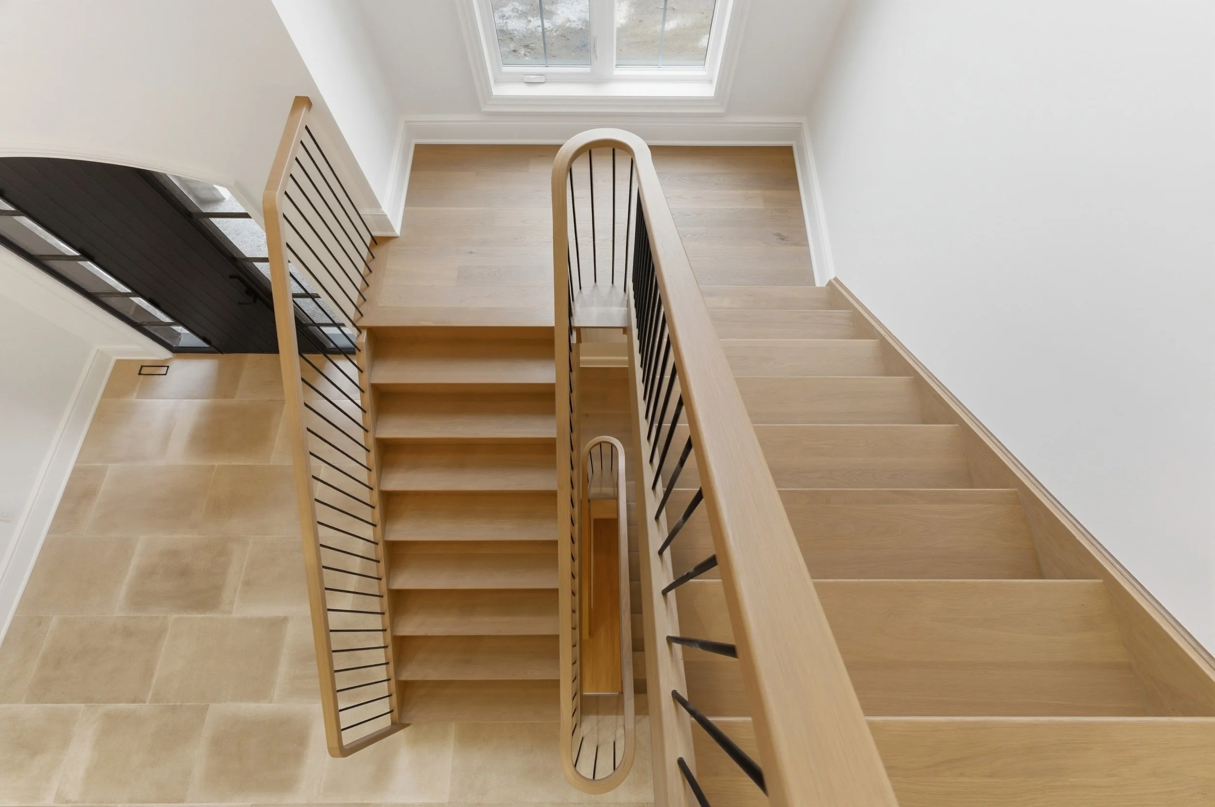 Top view of a modern wooden staircase with black railings, leading down to a tiled floor and a neighboring black door, onto a landing with a large window at the top.