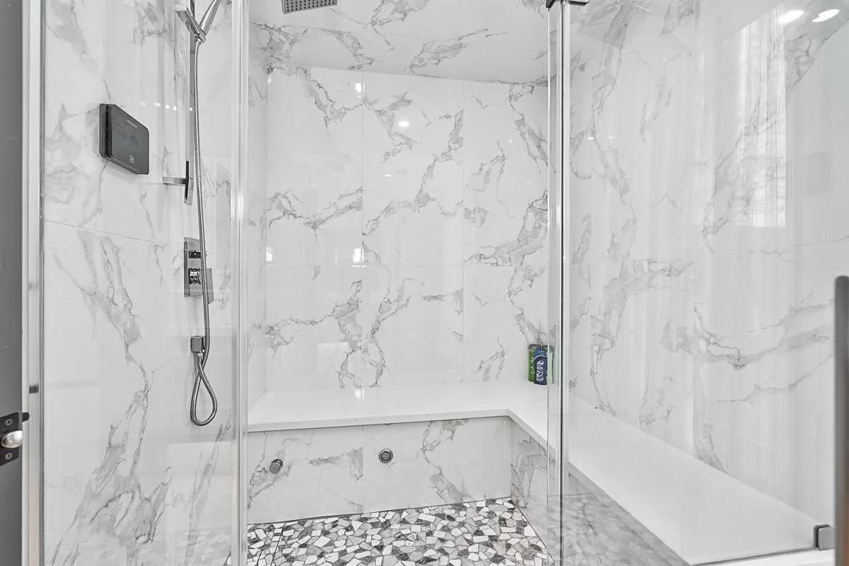 Modern walk-in shower with marble walls, a built-in bench, a rainfall showerhead, a handheld shower, and a pebble tile floor.