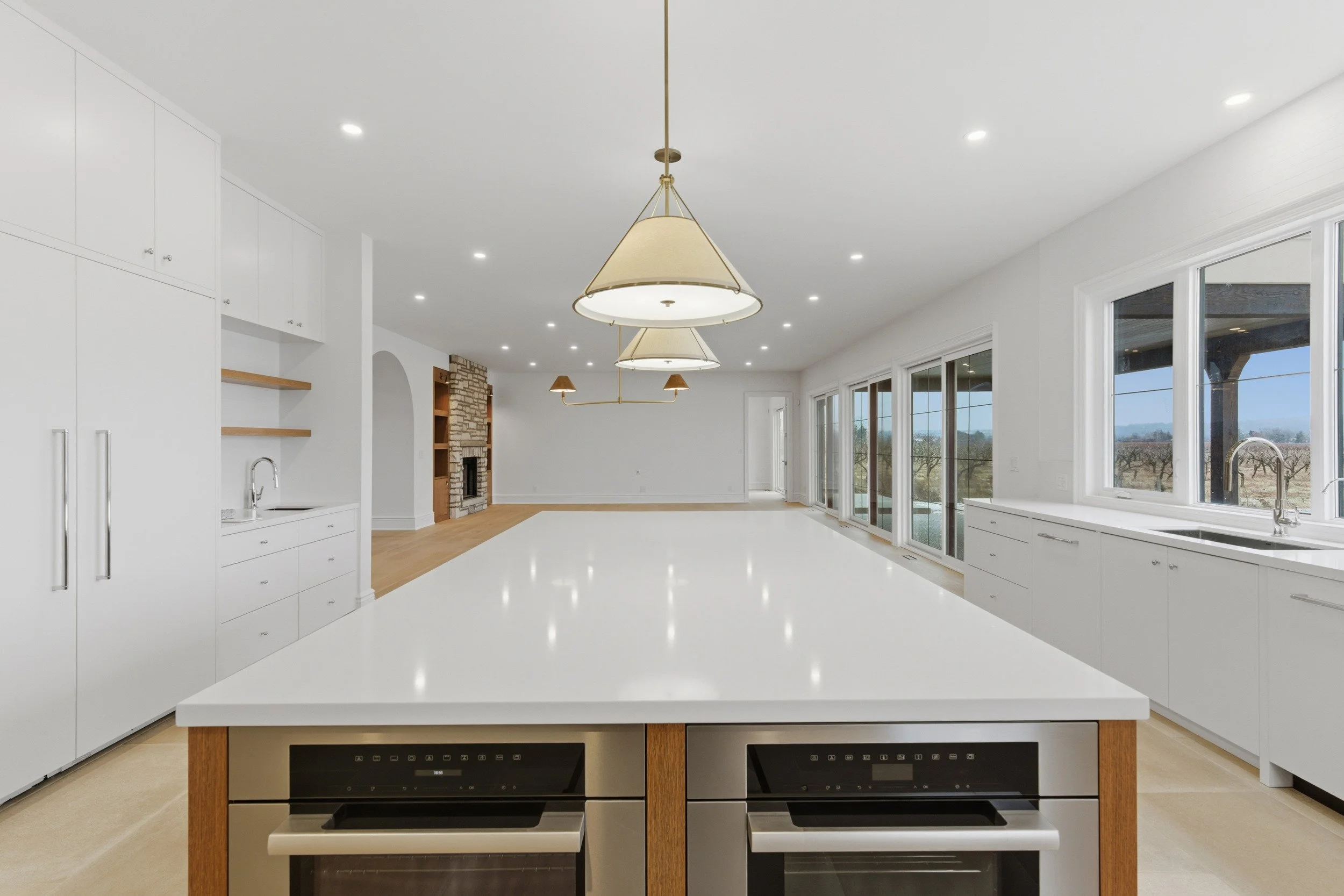 Modern kitchen with white cabinets, large island with white countertop, and plentiful natural light from windows. Pendant lights hang above the island, and a brick fireplace is visible in the background.
