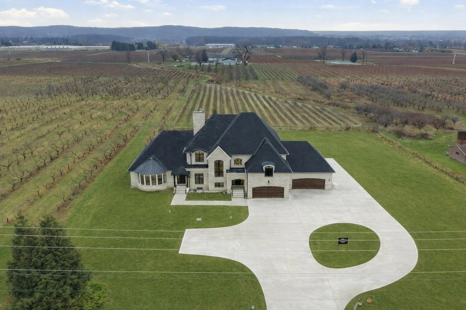 A large, elegant house with a dark roof and light exterior walls, surrounded by a spacious lawn and a circular driveway, in a rural area with vineyards and farmland in the background.
