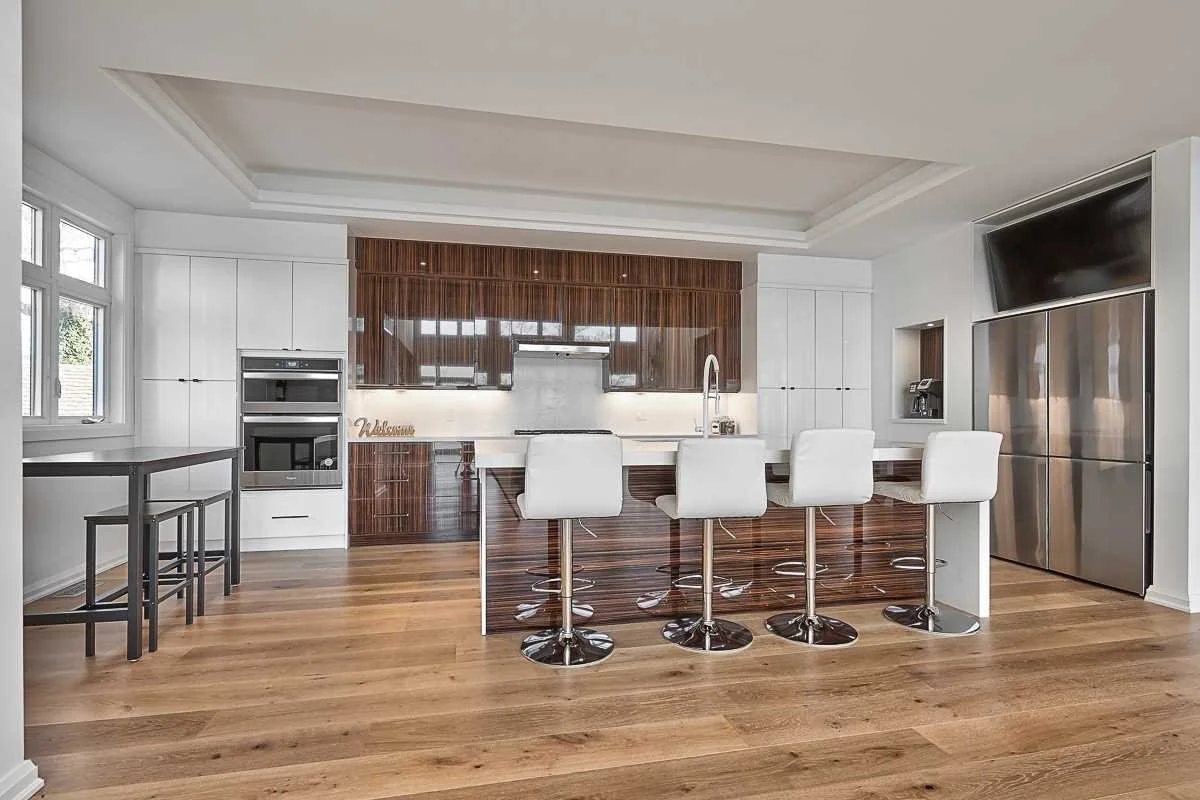 Modern kitchen with white and wood cabinets, four white bar stools at a marble island, stainless steel appliances, and a small table with two stools near a window.