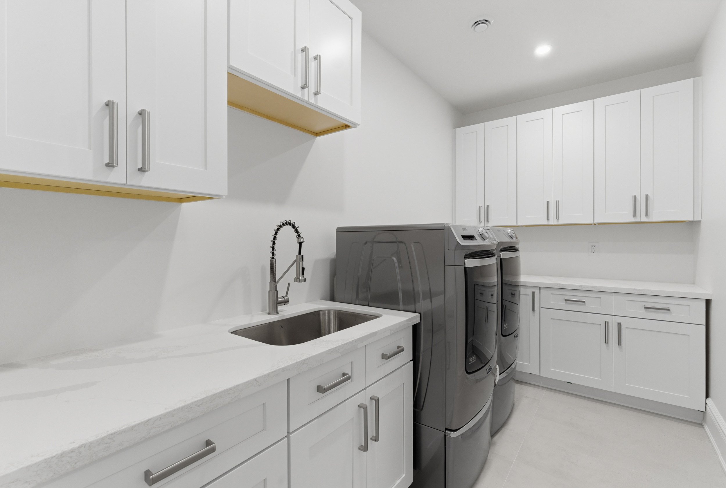 A laundry room with white cabinets, a white countertop, a stainless steel sink with a gooseneck faucet, and a pair of gray front-loading washing machines.