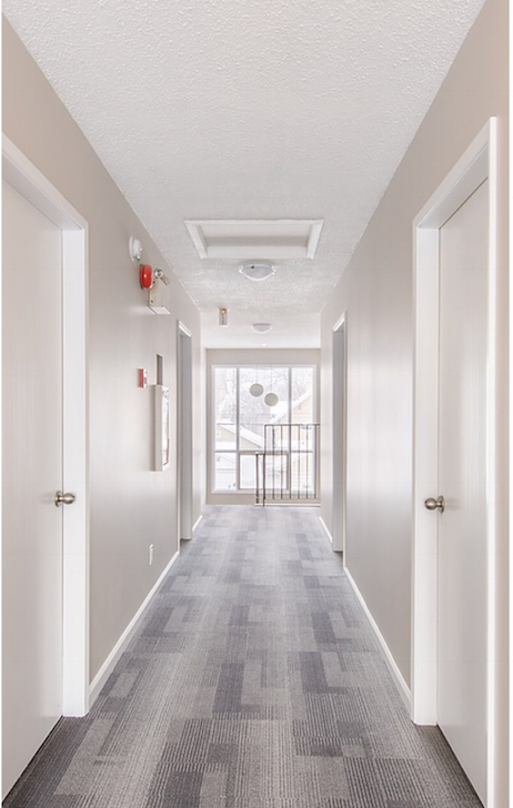 Empty apartment hallway with four closed white doors, beige walls, gray patterned carpet, ceiling lights, and a windowed door at the end allowing natural light.