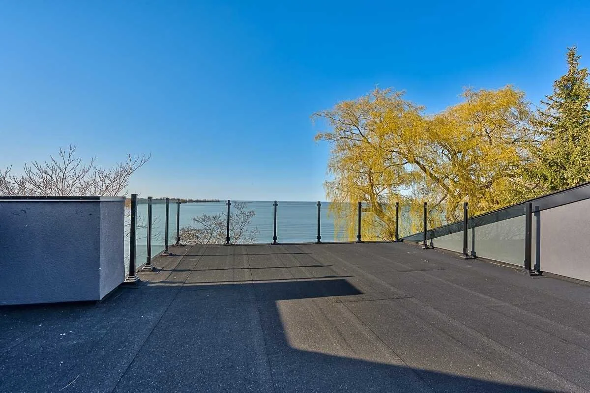 Rooftop terrace with glass railing overlooking water and trees, clear blue sky.
