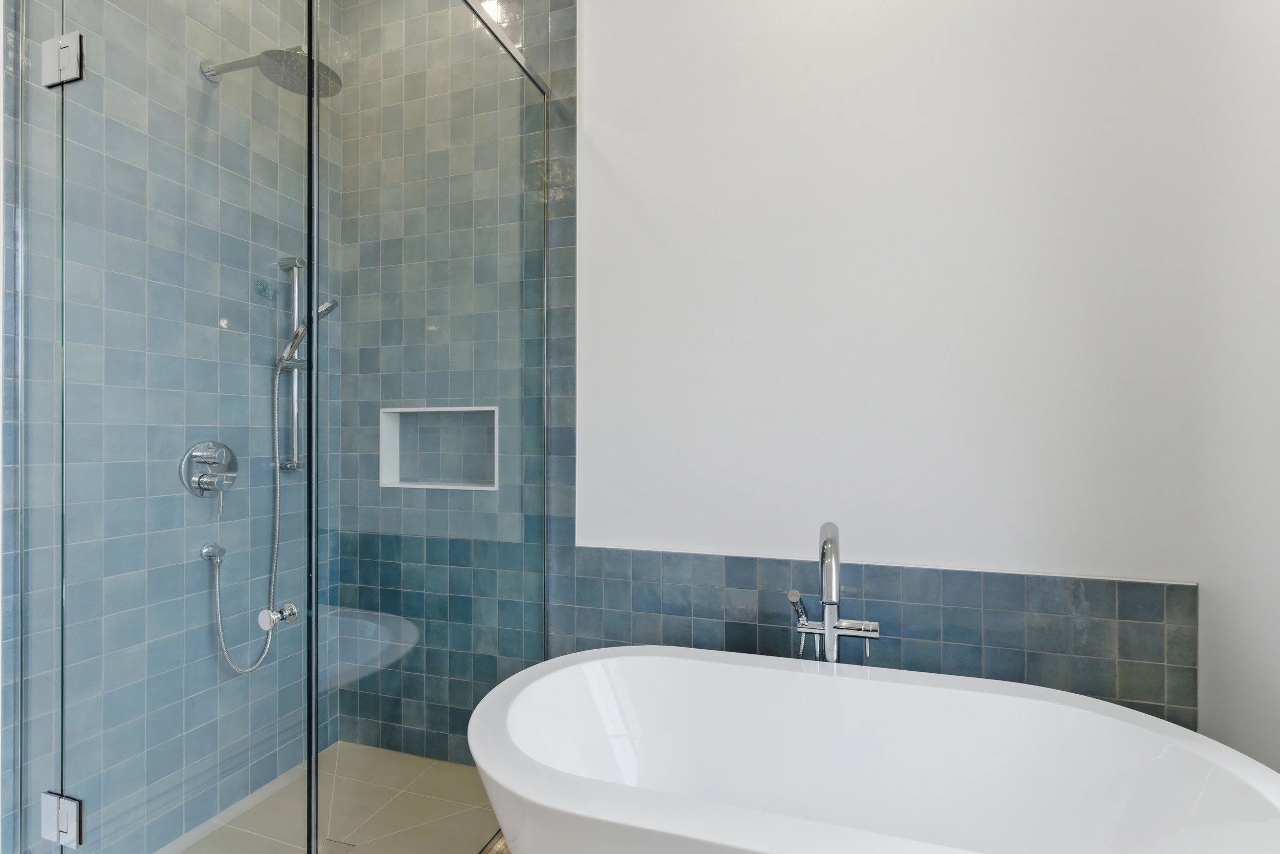 Modern bathroom with a glass-enclosed shower featuring water fixtures and a built-in shelf, adjacent to a white bathtub under a chrome faucet, with tiled walls and beige floor tiles.