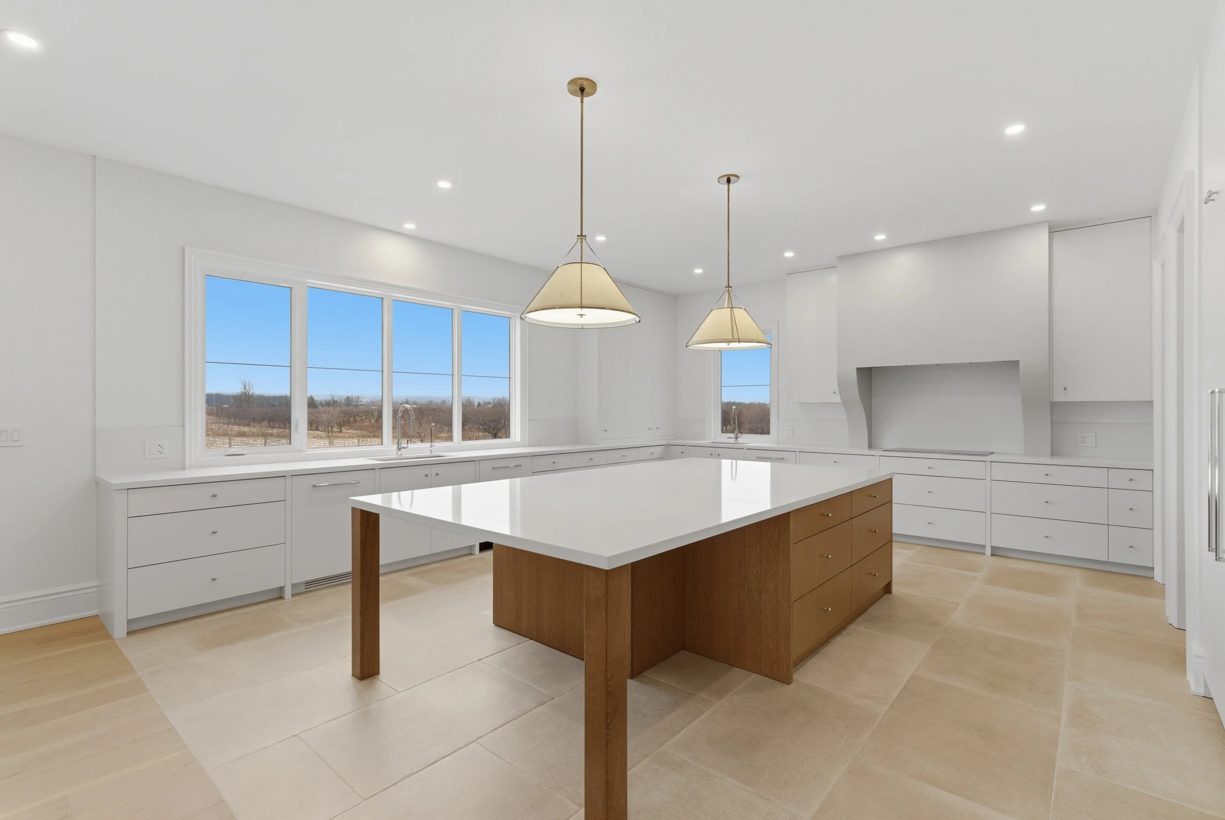 A modern white kitchen with large windows overlooking a rural landscape, featuring a white island with a wooden base, pendant lights, and white cabinetry.