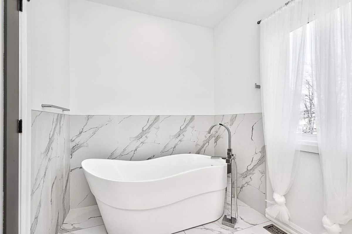 A white bathtub with a curved shape in a bathroom decorated with white marble tiles, next to a window with white curtains.