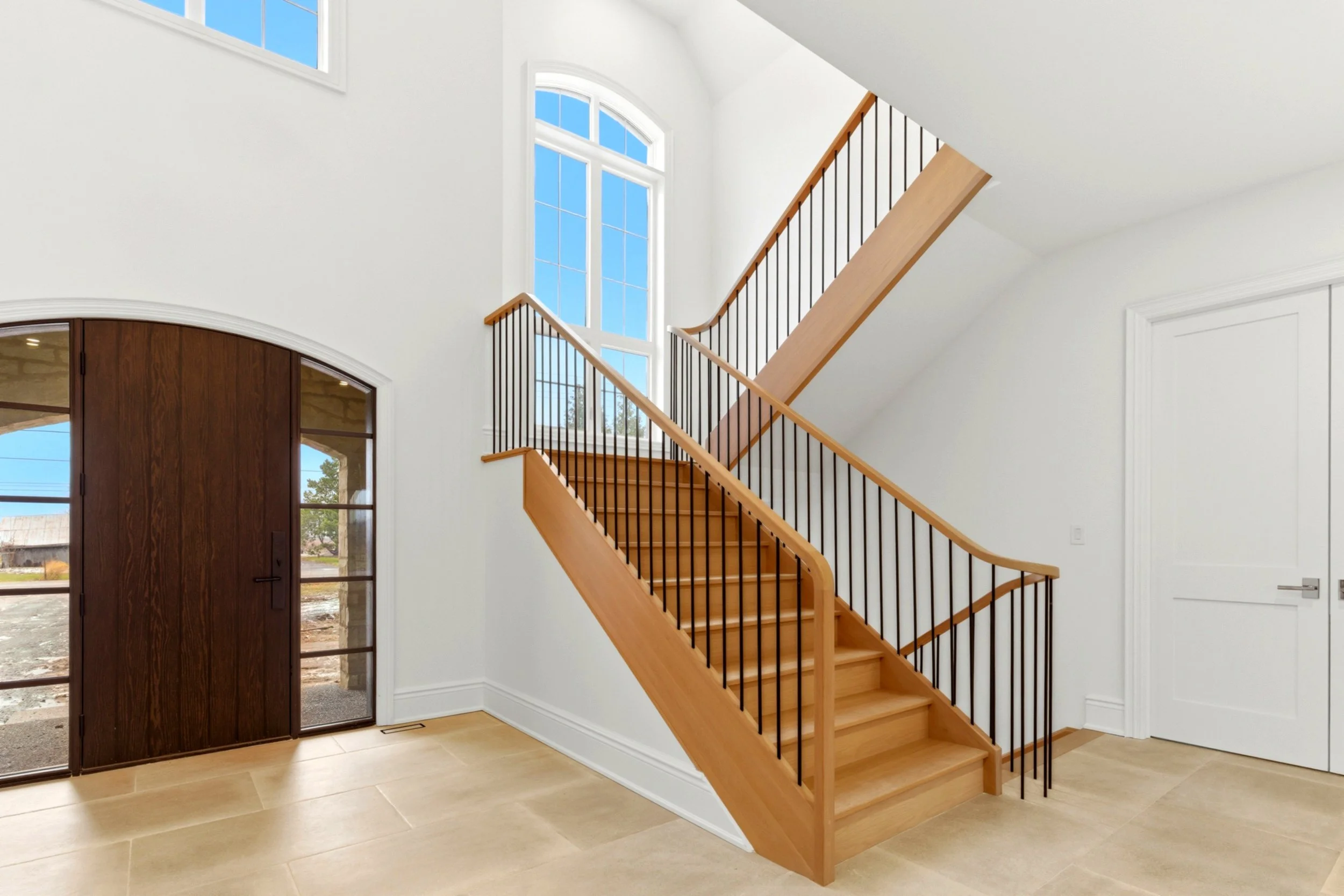 Interior view of a modern hallway featuring a wooden staircase with black metal balusters leading to the upper floor, large arched windows, a white wall, a white door, and a dark wood front door with glass panels.