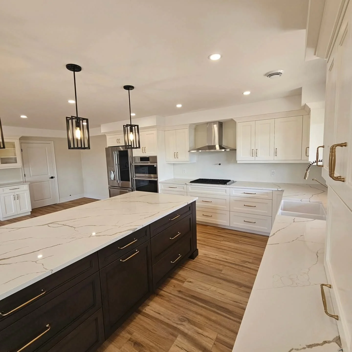 Modern kitchen with white cabinets, a dark central island with gold handles, marble countertops, and wooden flooring. There are two black pendant lights hanging from the ceiling, and stainless steel appliances including a refrigerator, oven, microwav