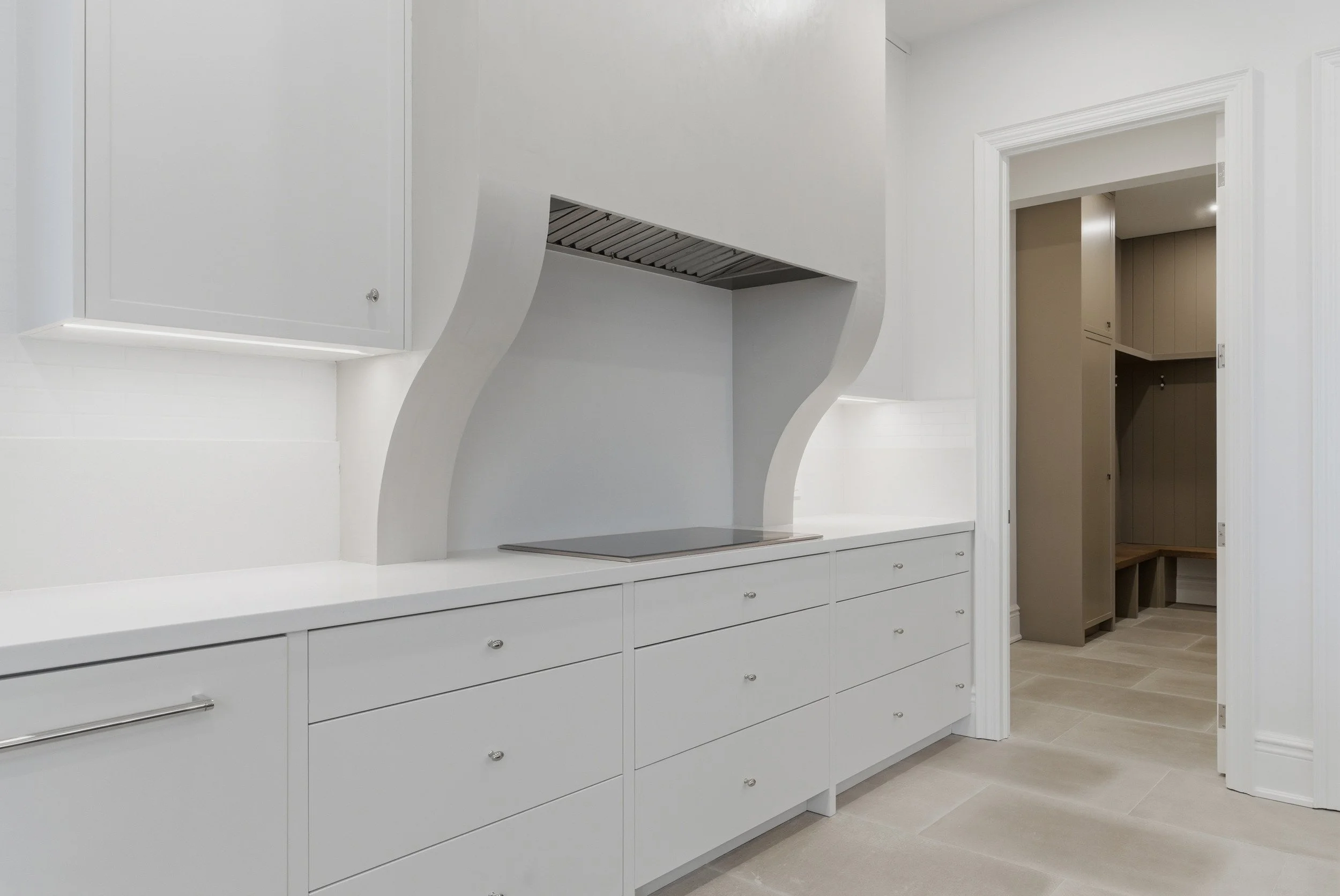 White kitchen with built-in cabinetry, smooth countertop, and a cooktop under a modern range hood with a curved design. Adjacent open pantry with seating area.