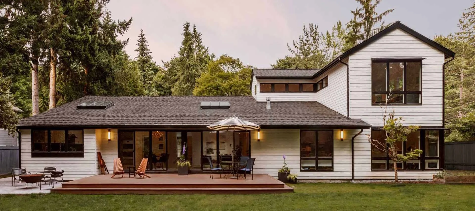 Modern two-story house with a deck, surrounded by trees, featuring white siding, large windows, a patio umbrella, and outdoor seating.