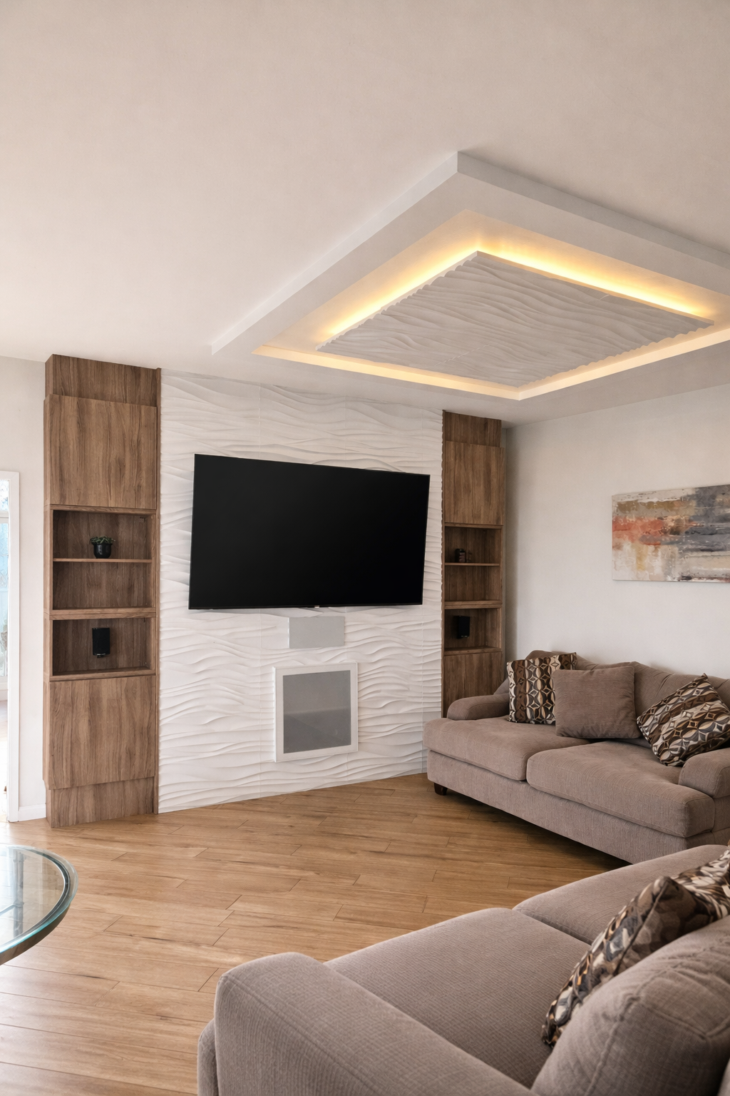 Living room with beige sofas, patterned pillows, a wall-mounted TV, wooden flooring, and built-in wooden shelves.