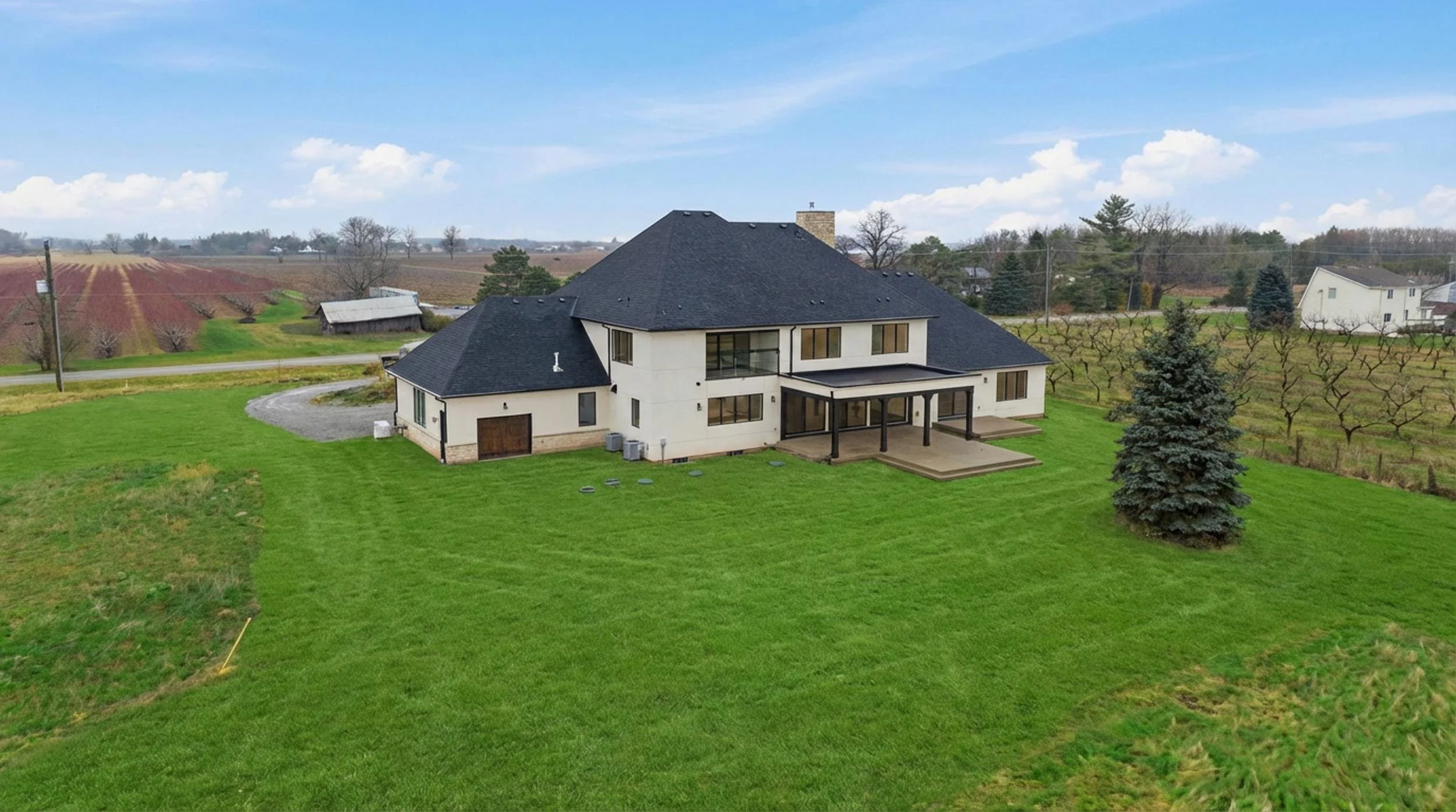 A large modern house with a dark roof and white exterior walls, surrounded by a green lawn and trees, with farmland and other houses in the background under a partly cloudy sky.
