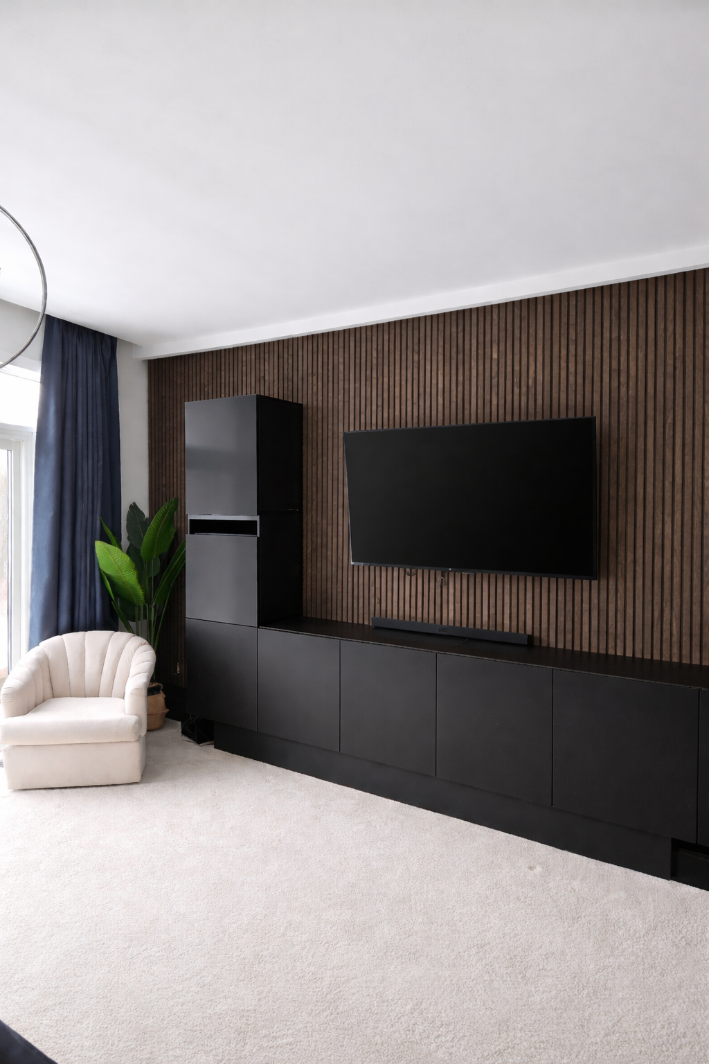 Modern living room with a wall-mounted flat screen TV, black entertainment unit, plant, armchair, and large window with blue curtains.