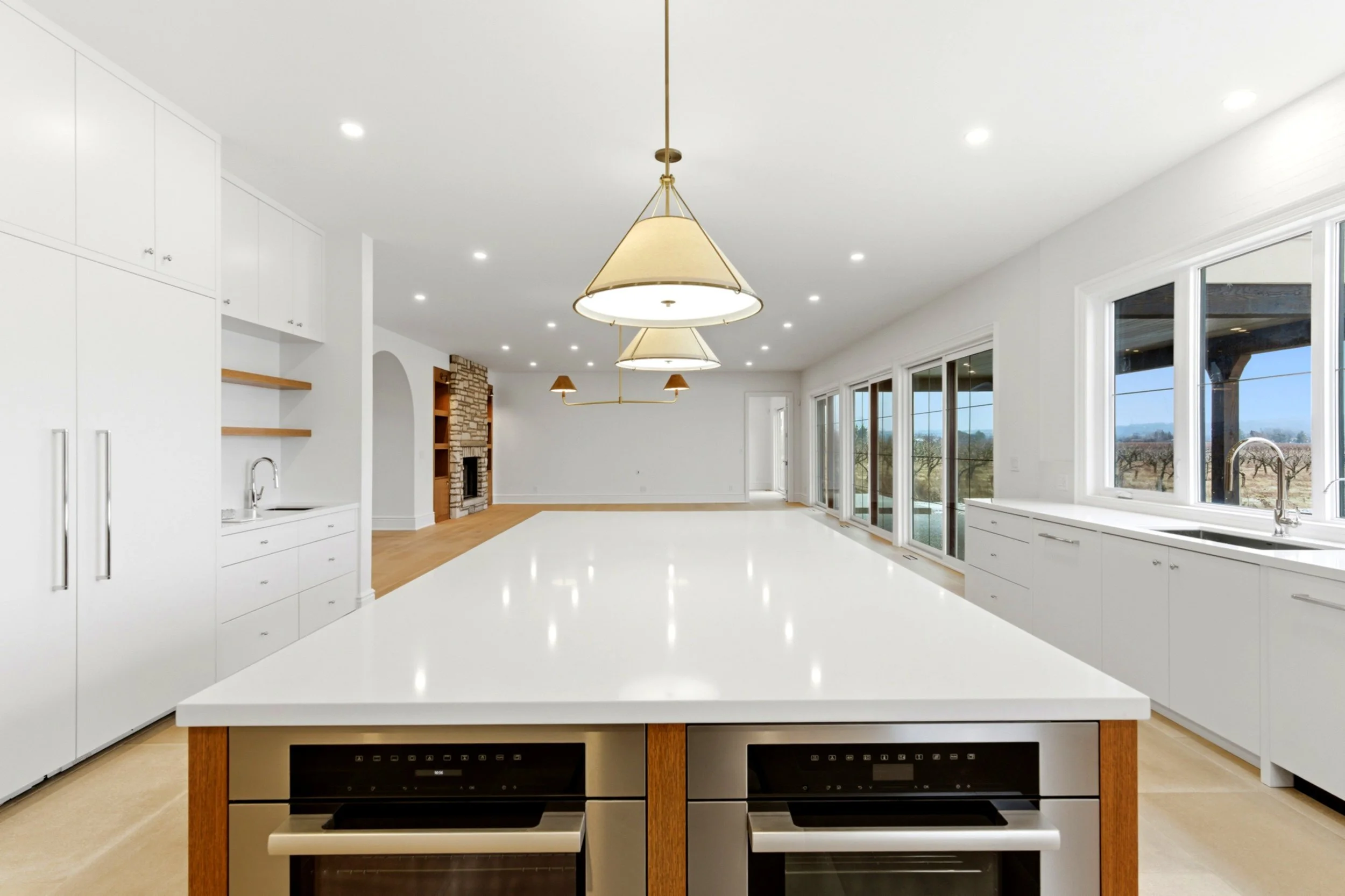 Bright, spacious kitchen with white cabinets, a large white countertop, and windows overlooking a backyard. There is a double oven under the island and a sink beneath the windows. The room opens to a living area with a stone fireplace and views of the outdoors.