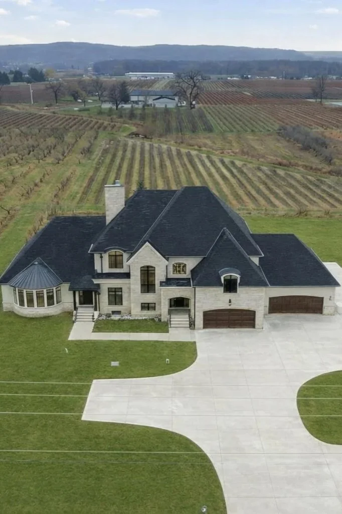 Large modern house with black roof and light exterior, paved driveway, well-kept lawn, situated on expansive farmland with vineyards and distant hills.