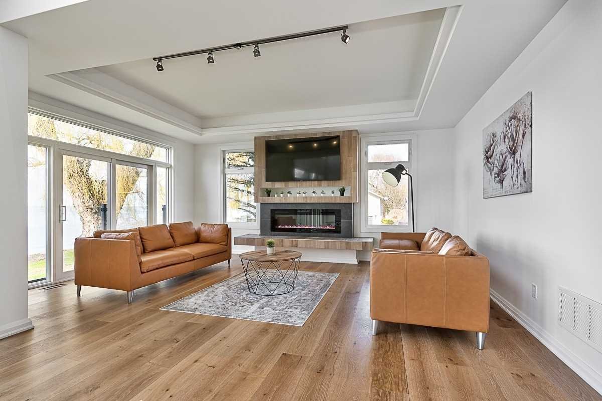 Modern living room with light wood floors, two tan leather sofas, a round coffee table, a wall-mounted TV, a fireplace, large windows, and a floor lamp.