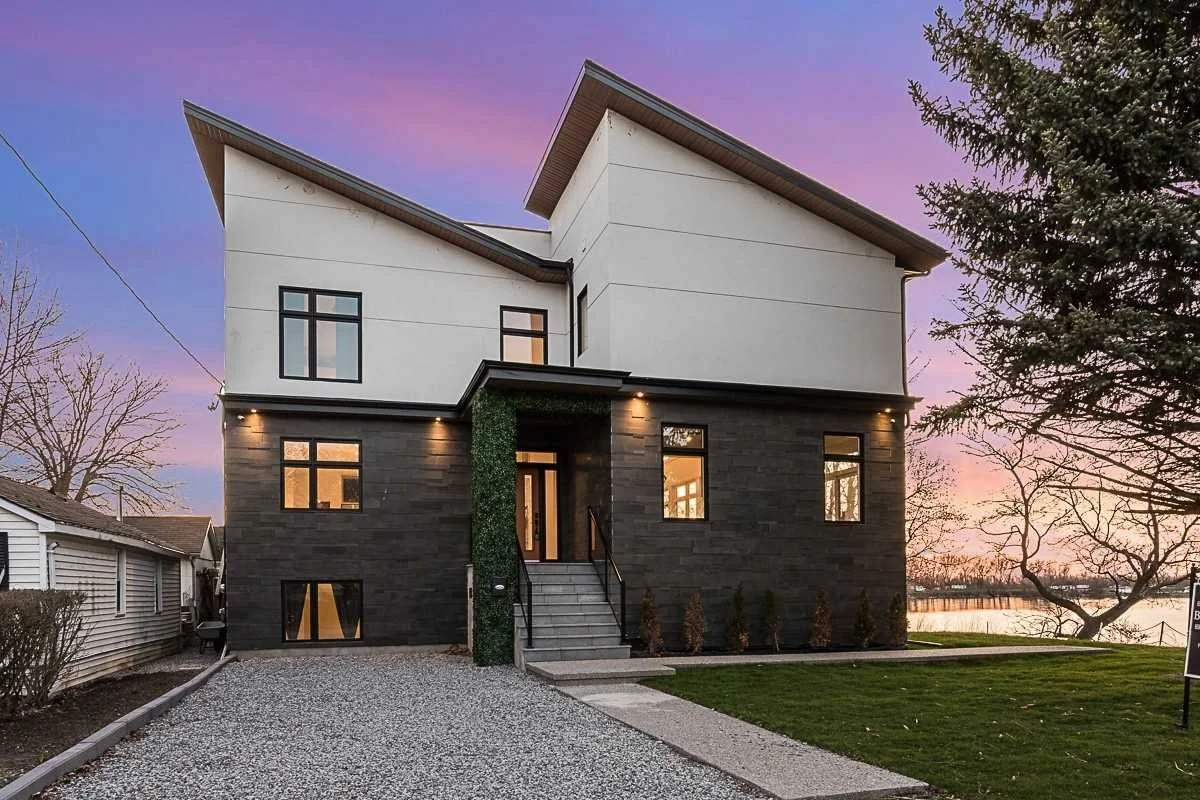 Modern two-story house with a white upper floor and black lower floor, set against a sunset sky with purple and pink hues. The house has a gravel driveway, small front steps, and a lush lawn with a few plants at the entrance. Trees and water are visi