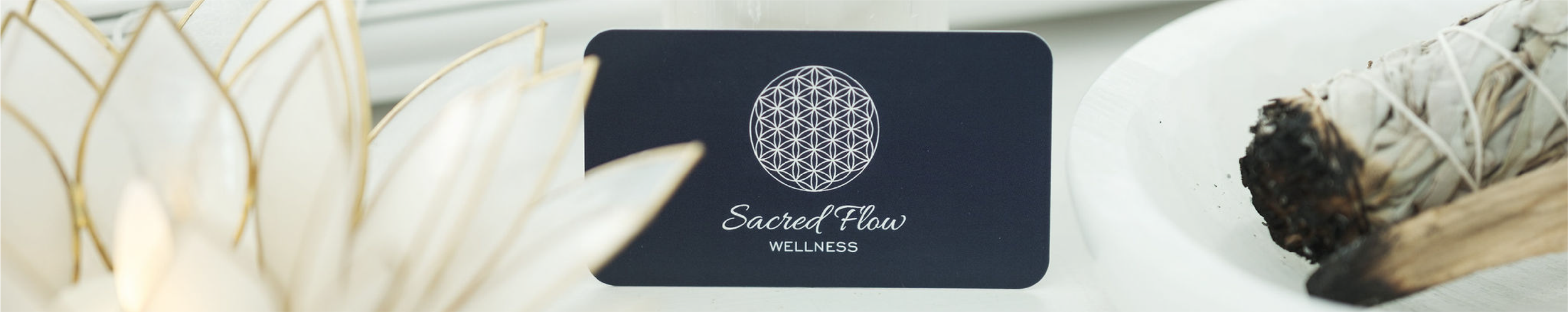 A black card with a geometric flower of life design and the text 'Sacred Flow Wellness' placed on a white surface, surrounded by white and beige decorative elements.