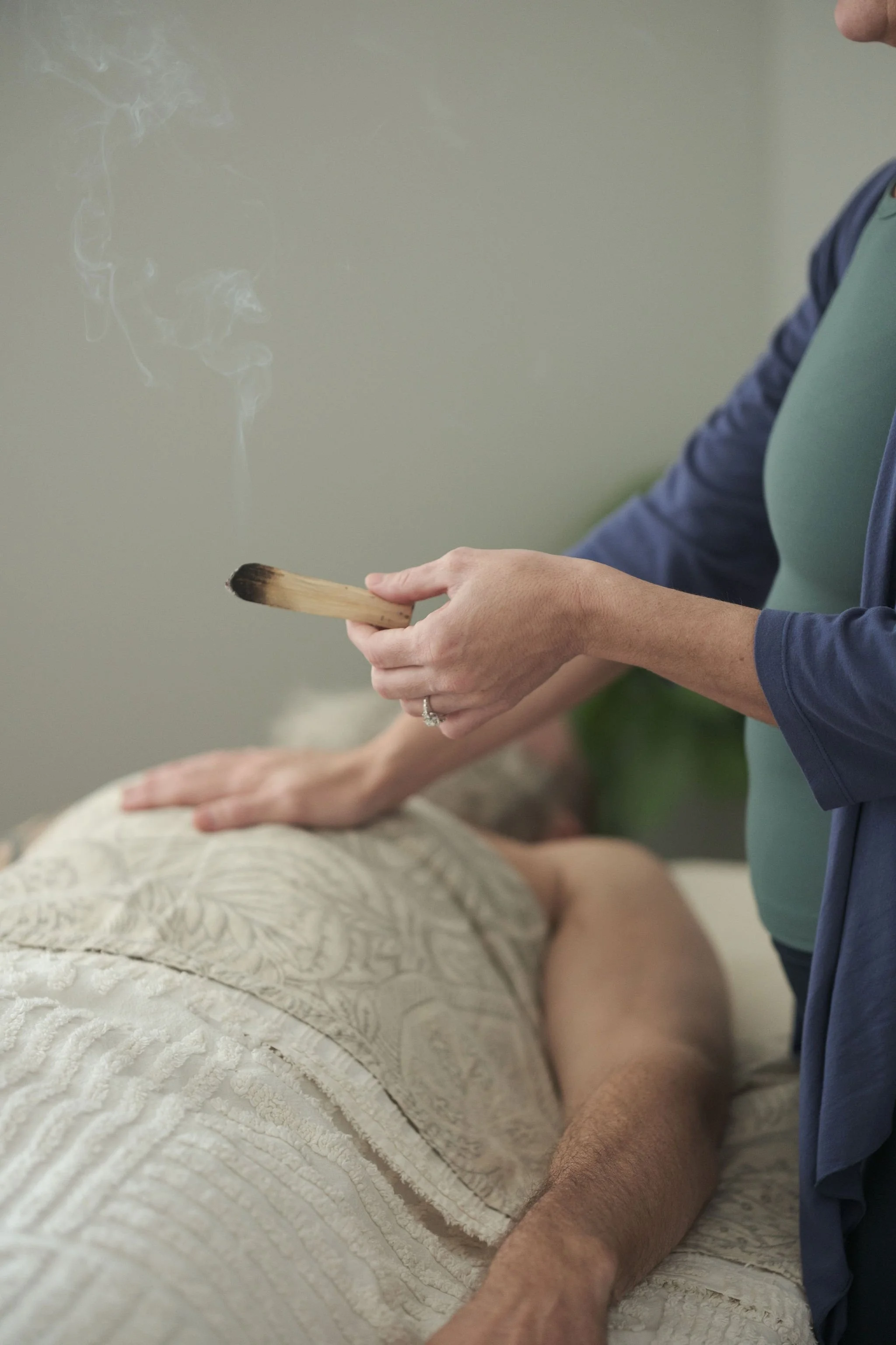 Person holding smudging stick over someone lying face down on a bed, with a cloud of smoke rising in a ceremony or ritual.