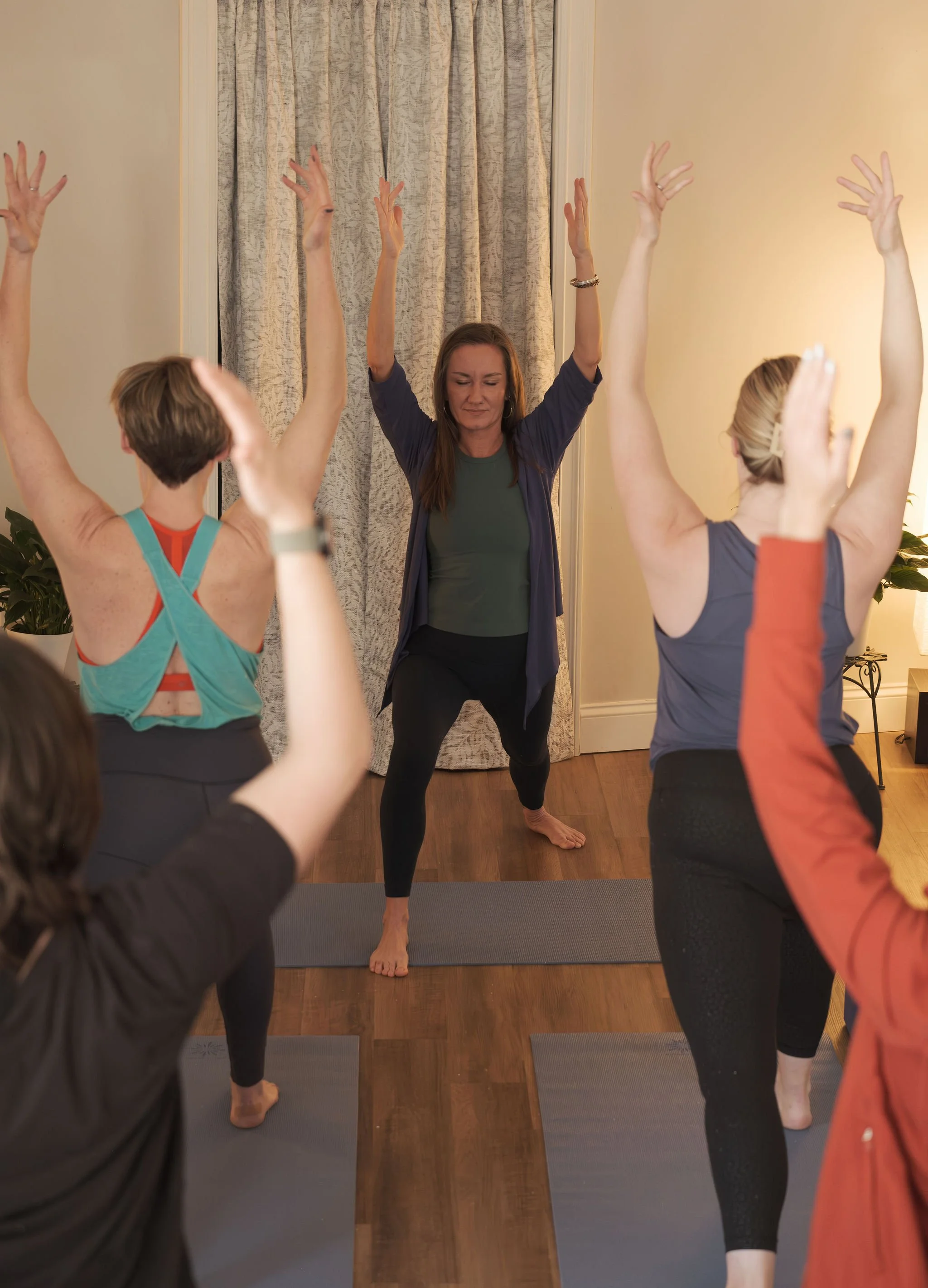 A woman leading a yoga class with her eyes closed, practicing a tree pose with one foot on the mat and arms raised, surrounded by participants also doing the pose.