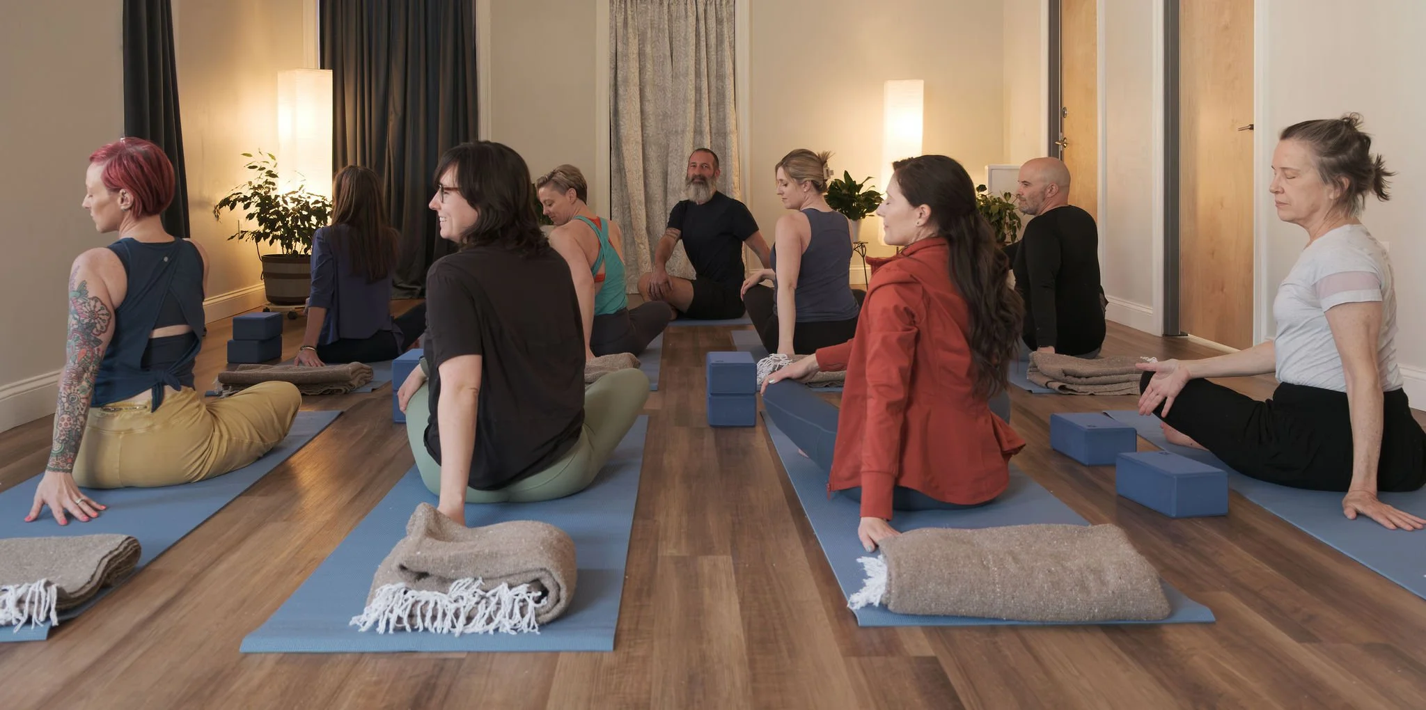 A group of people practicing yoga together in a room, sitting on yoga mats with folded towels and yoga blocks nearby, with plants and warm lighting in the background.