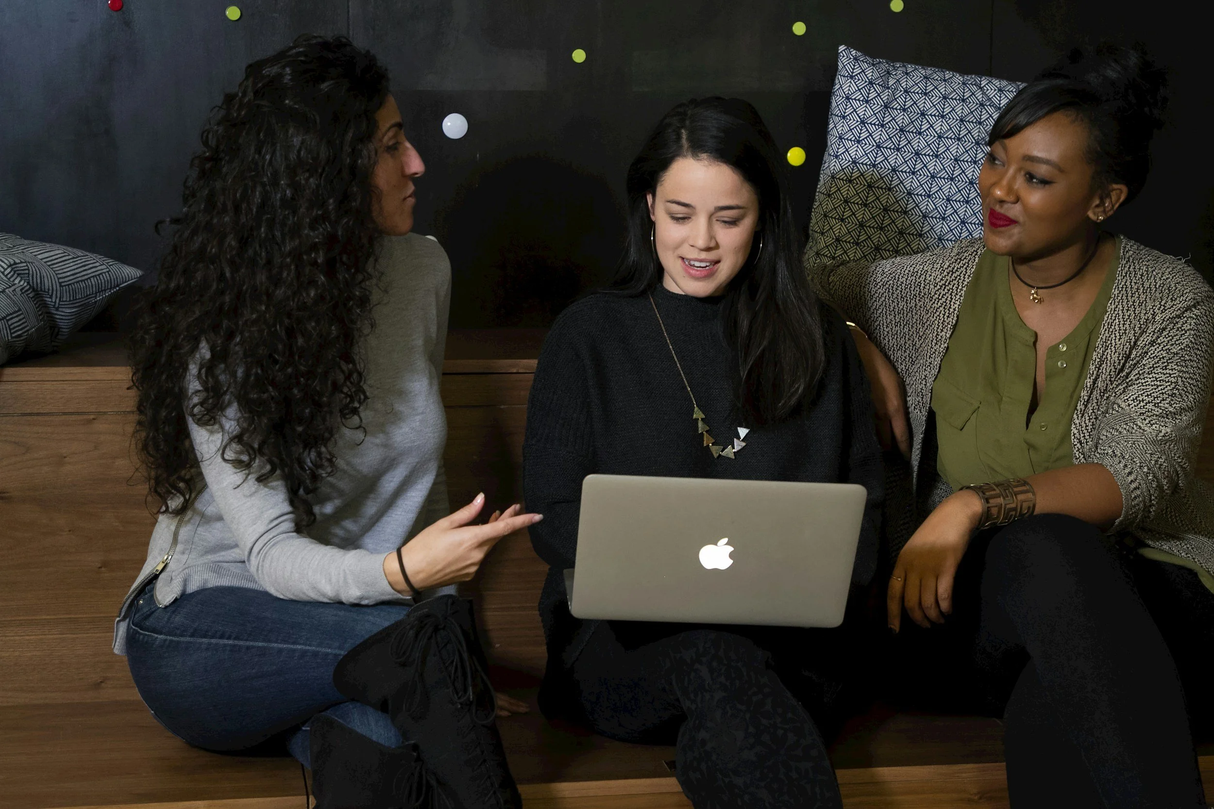 Three women sitting closely together on the floor, looking at a laptop screen, in a cozy, modern room with dark walls and decorative pillows.