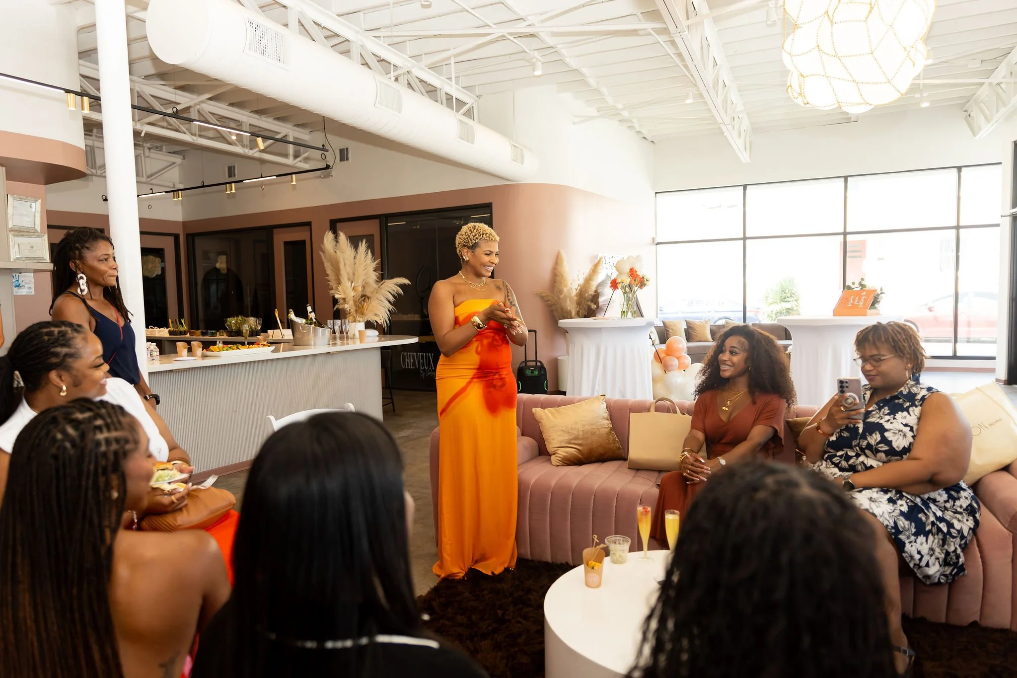 A group of women gathered in a modern, stylish cafe or lounge, with one woman standing and speaking to the others, dressed in a vibrant orange and yellow dress. The women are seated on a pink sofa and around a white coffee table, some holding drinks and one taking a picture. The space has large windows, modern decor, and a light color scheme.