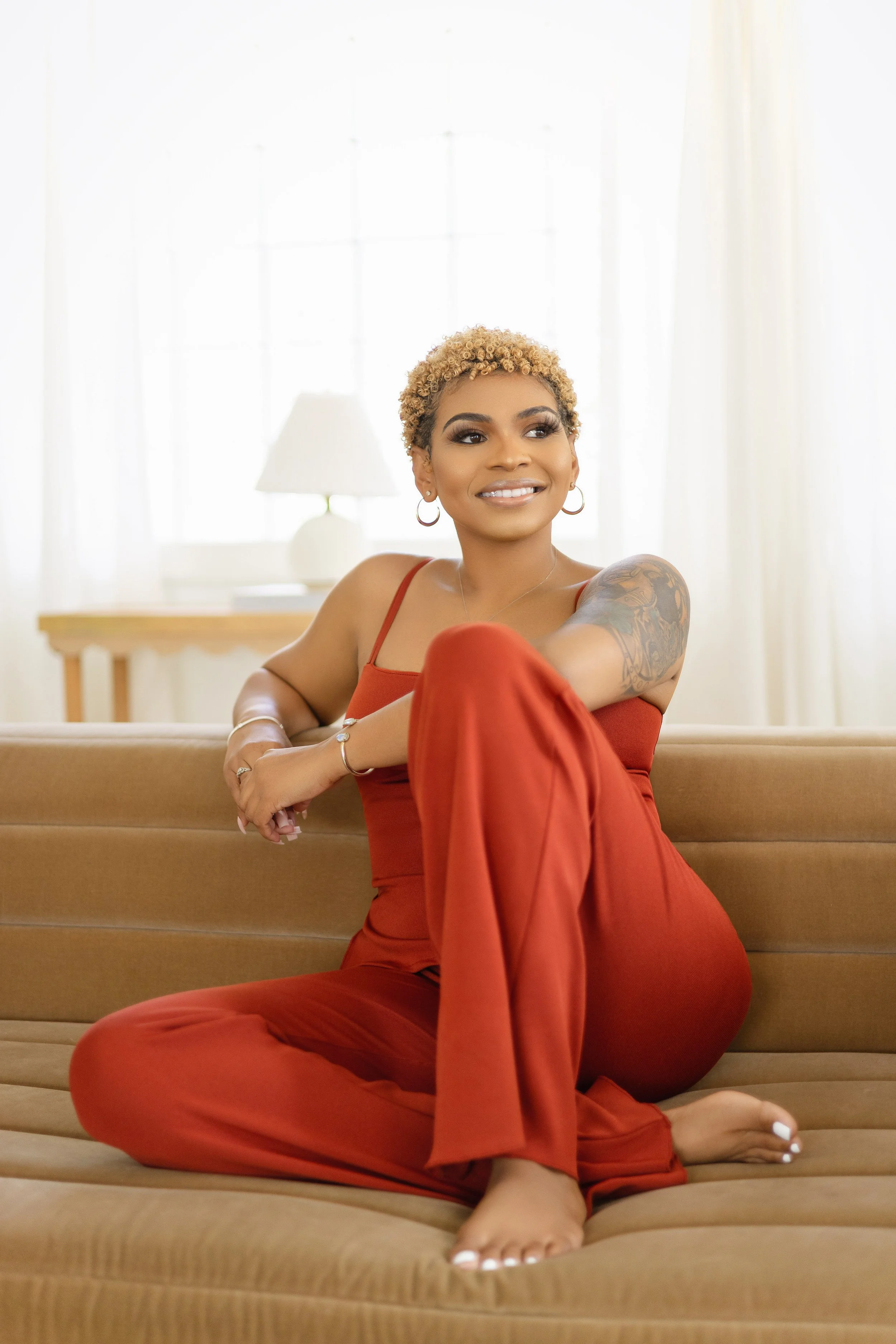 A woman with short curly blond hair, wearing a red jumpsuit, sitting on a beige sofa in a brightly lit room with white curtains and a lamp in the background, smiling and looking to her left.