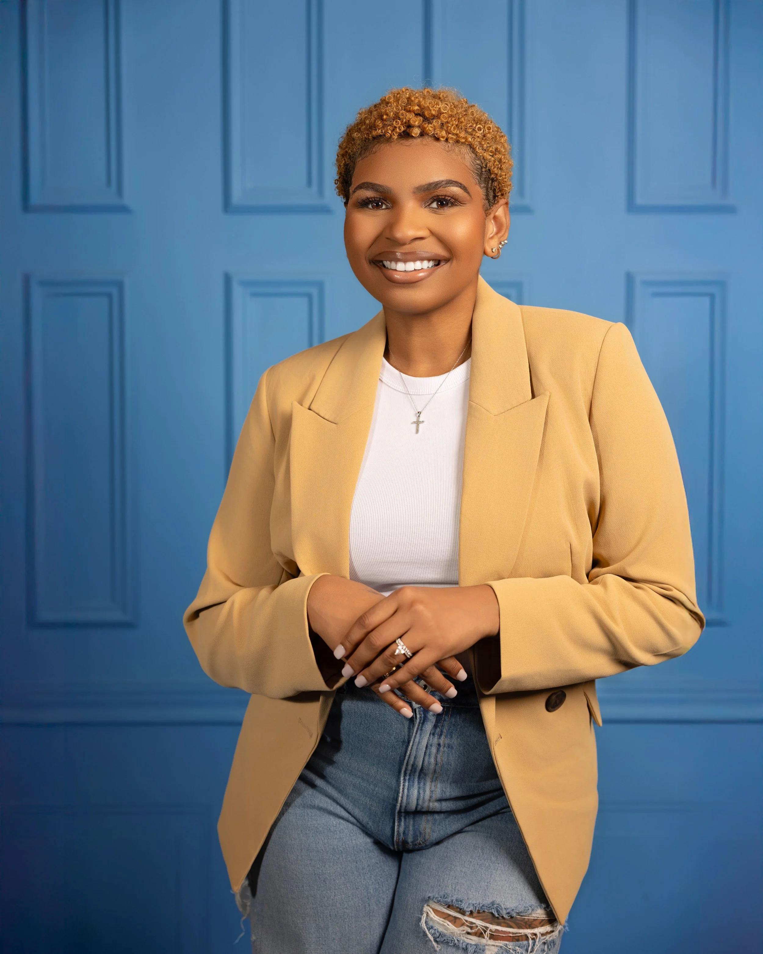 A woman with short curly blonde hair smiling, wearing a tan blazer over a white top, blue jeans with a tear, standing in front of a blue paneled wall.