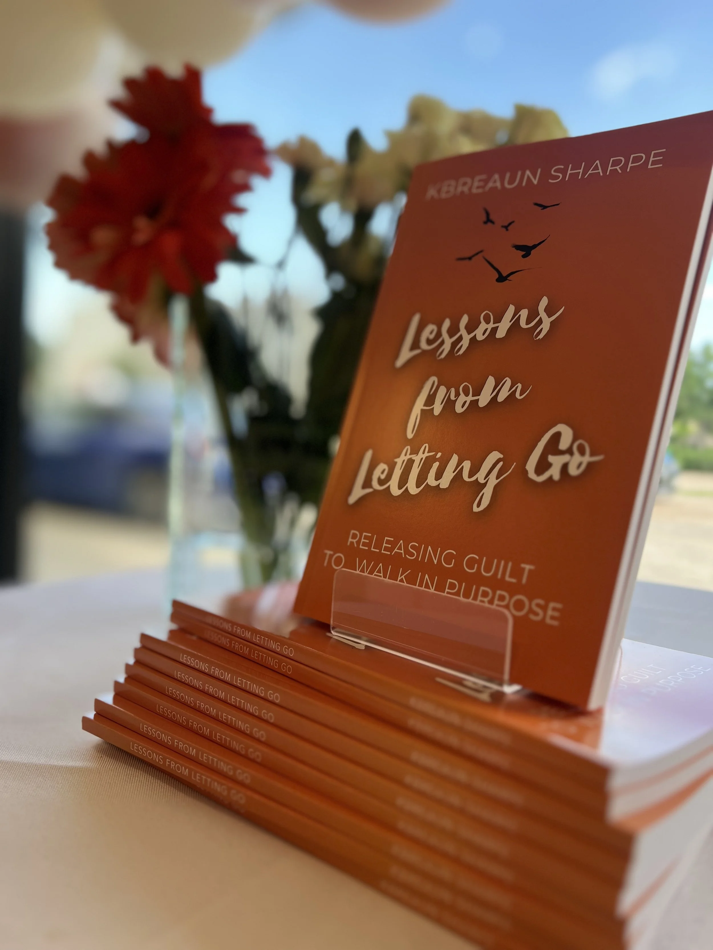 Stack of orange books titled "Lessons from Letting Go" by Kbreaun Sharpe, with a vase of red and white flowers in the background.