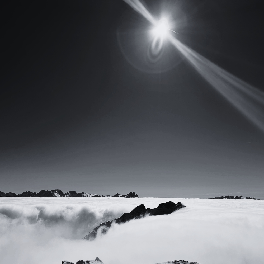 Un paysage en noir et blanc avec des rochers émergeant d'une mer de nuages, un ciel clair et un soleil brillant créant un phénomène de halo lumineux.