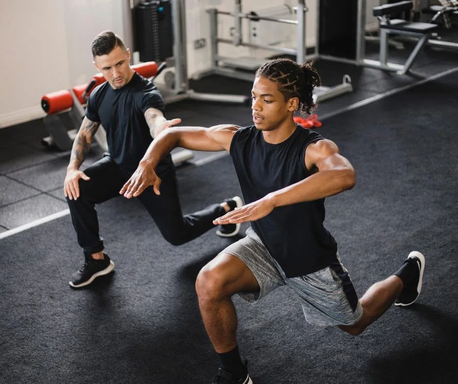 A personal trainer assists a young man with a seated lunge exercise in a gym.