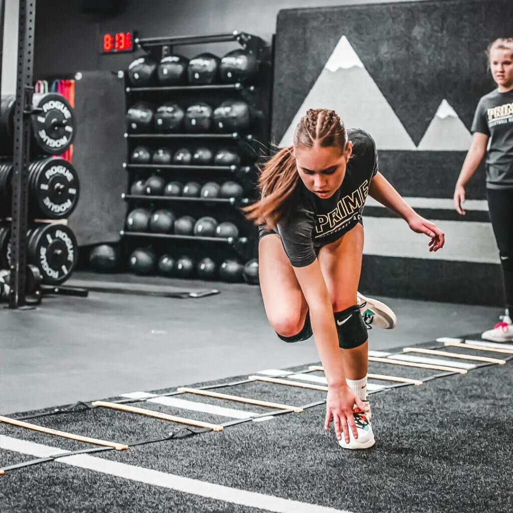 Young woman performing a balancing exercise on agility ladders at a gym with black weights and kettlebells in the background.