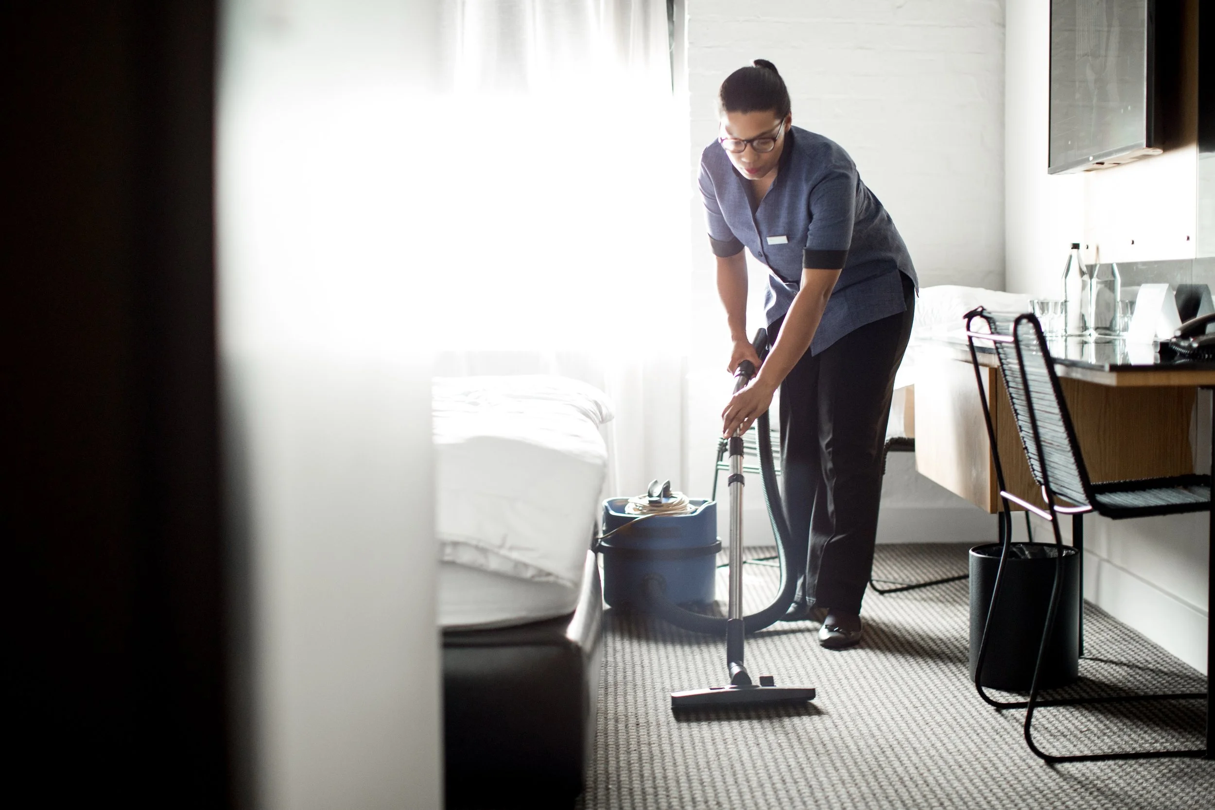A woman wearing glasses and a blue uniform vacuuming a hotel room with a portable vacuum cleaner.