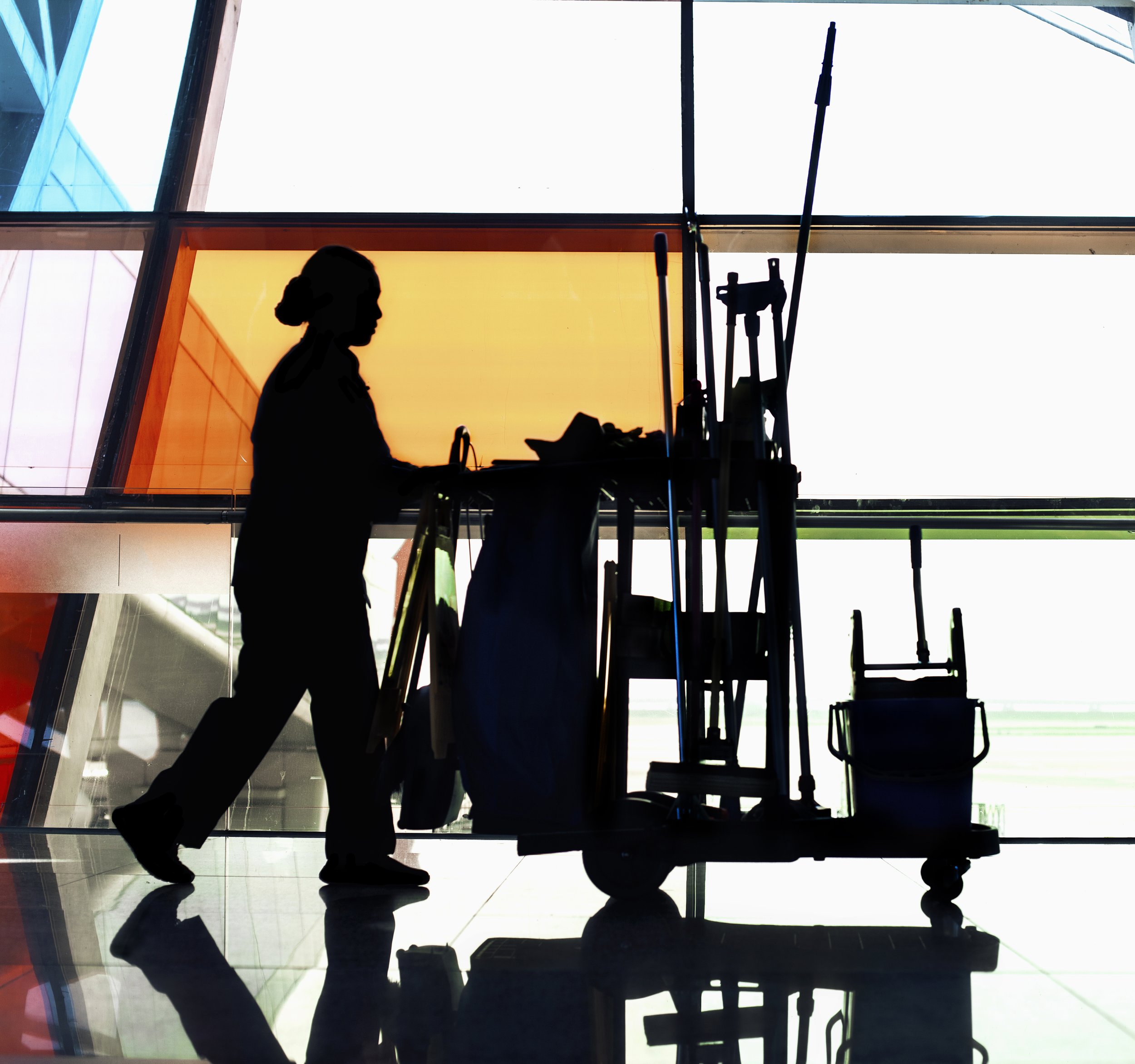 Silhouette of a person with a bun hairstyle standing near a cart or trolley filled with items, inside a building with large colorful windows showing blue, orange, and white sections, possibly at an airport or shopping mall, with reflections on the floor.
