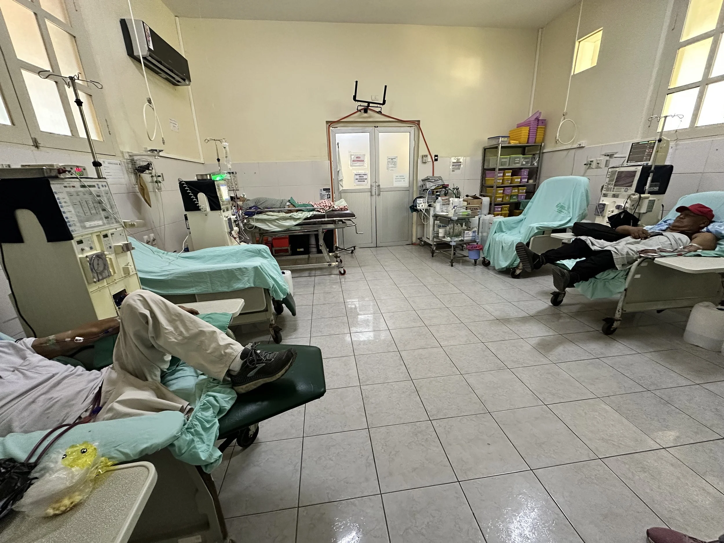 Hospital room with four patients lying on beds connected to medical equipment, windows on the right side, and a door at the back with medical supplies and equipment around.