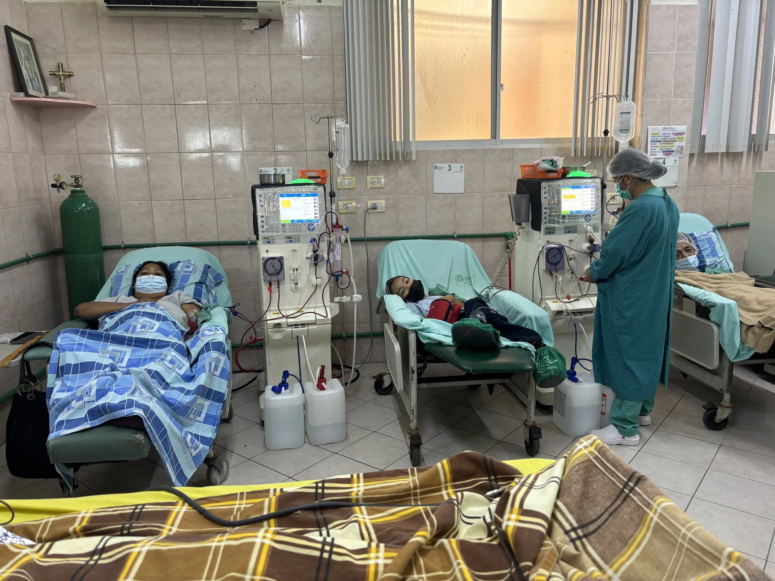 Hospital room with three patients lying on beds, all wearing masks. Medical staff in scrubs and masks attending to patients. Medical equipment and machines connected to patients, with supplies on the beds.
