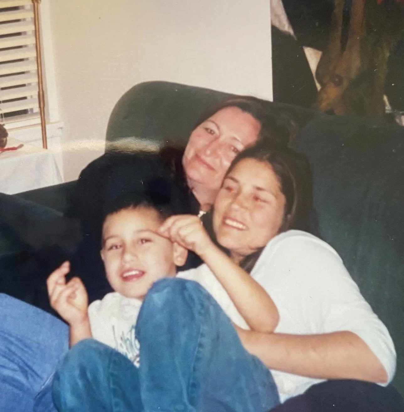 Three people cuddling on a dark couch, smiling, with a young boy in front, a woman in the middle, and a woman behind.