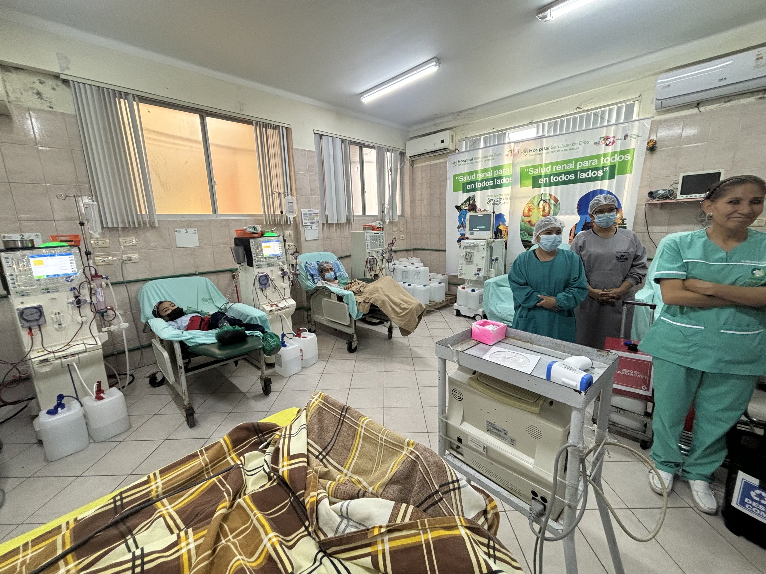 Hospital dialysis center with two patients on dialysis machines, including medical staff observing, in a room with tiled walls and medical equipment.