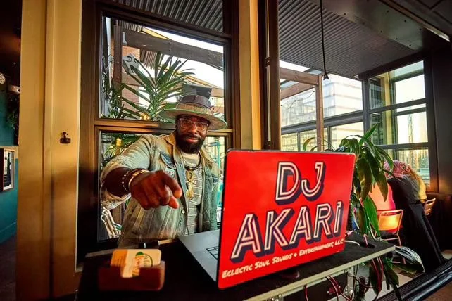 Man wearing a hat and glasses pointing at the camera behind a DJ booth with a red sign that says 'DJ AKARI'.