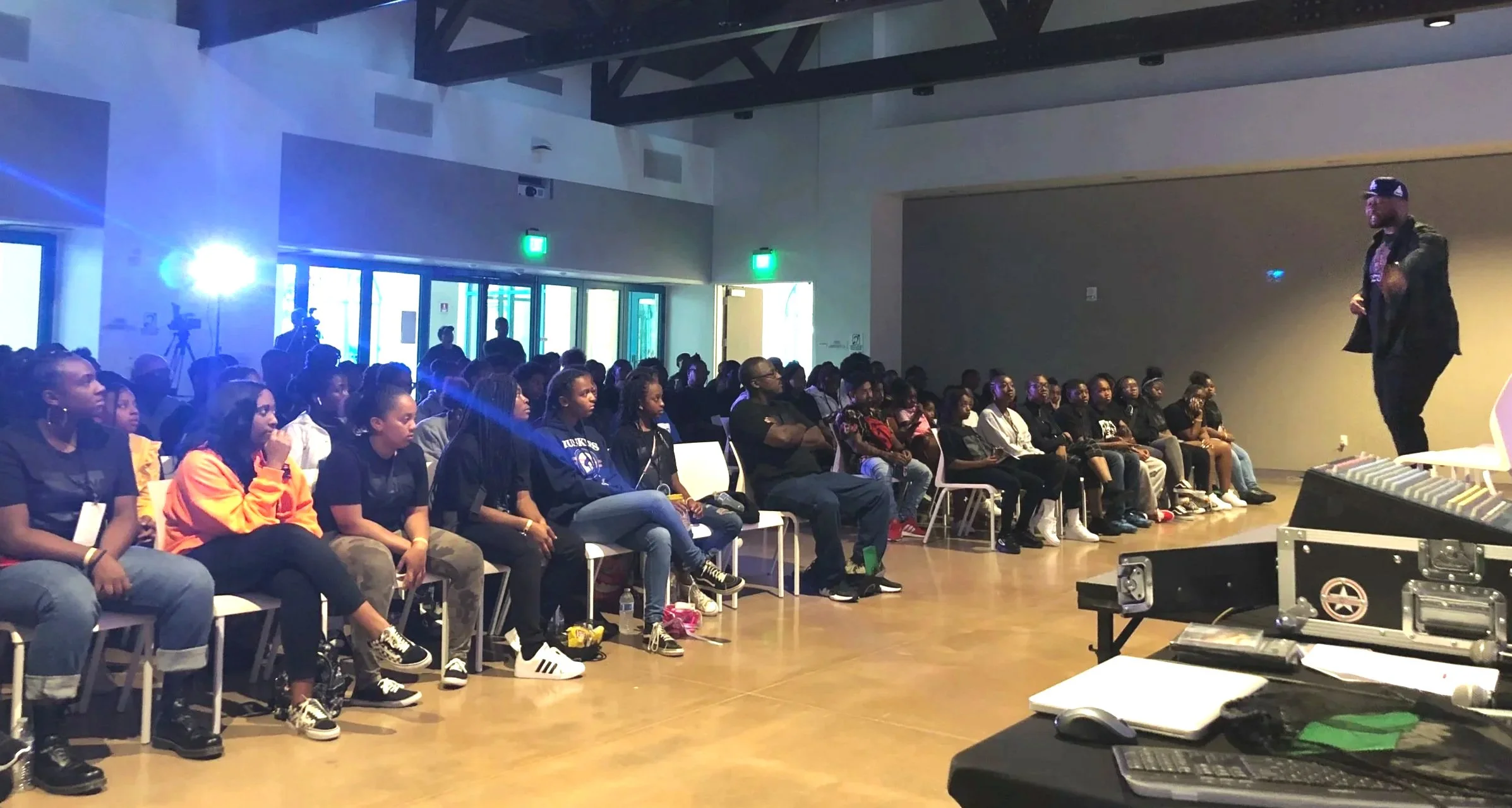 A speaker is on stage addressing a seated audience in a dimly lit room with blue and green lighting effects and camera crew recording the event.