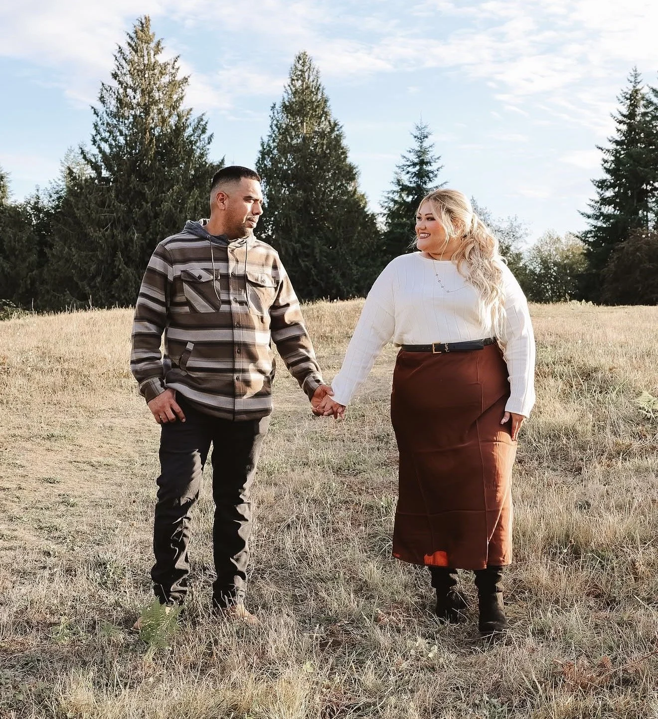A man and woman holding hands and standing in a grassy field with trees in the background. The man is wearing a striped hoodie and dark pants, while the woman is wearing a white sweater and a brown skirt.