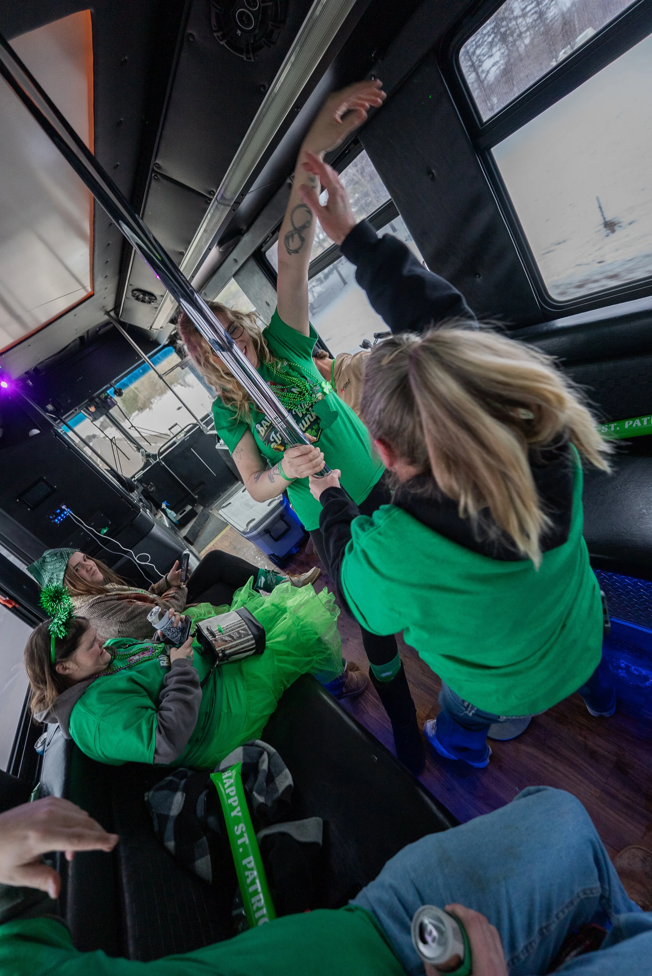 People inside a bus wearing green clothing and accessories, celebrating St. Patrick's Day, with some holding drinks and the bus decorated with green items.