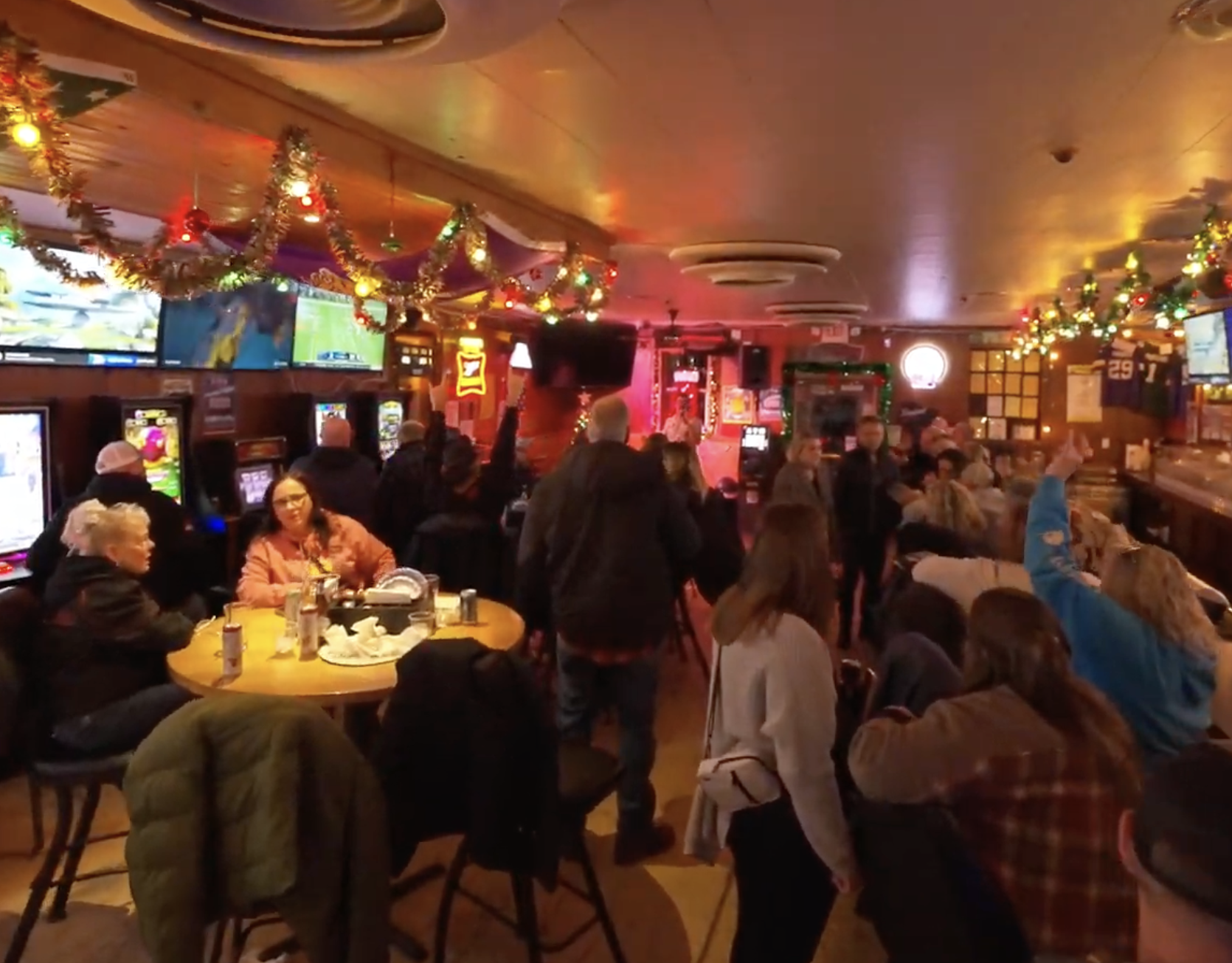 A lively bar decorated with Christmas tinsel, lights, and garlands, filled with people watching TV screens, sitting at tables, and socializing.