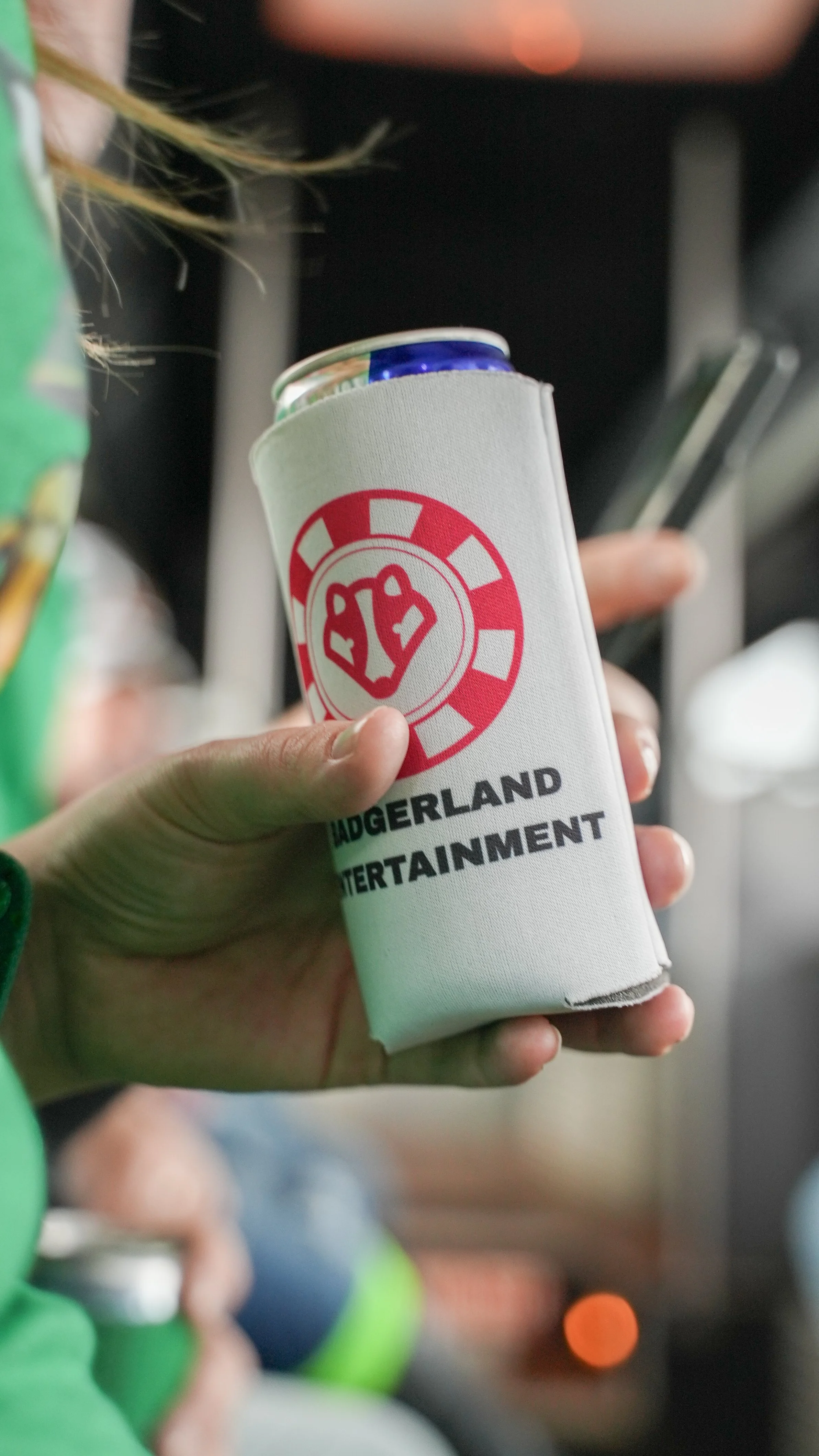A close-up of a hand holding a can cooler with a red and white logo and black text that reads 'HADGERLAND ENTERTAINMENT', with a can inside it. The background shows part of other cans and blurred objects.